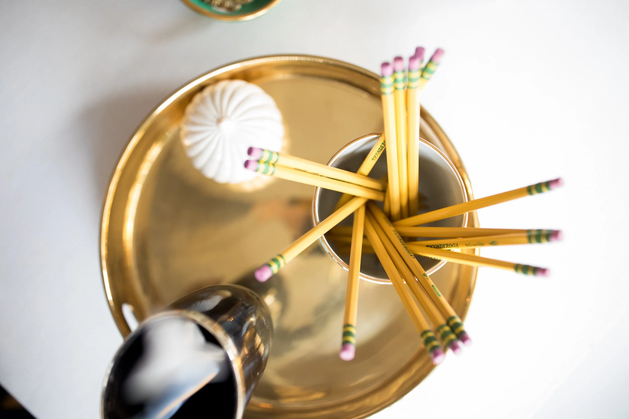 A golden tray holding black and white empty shot glasses and a glass with yellow pencils with pink eraser tips and green-striped bands, seen from above.