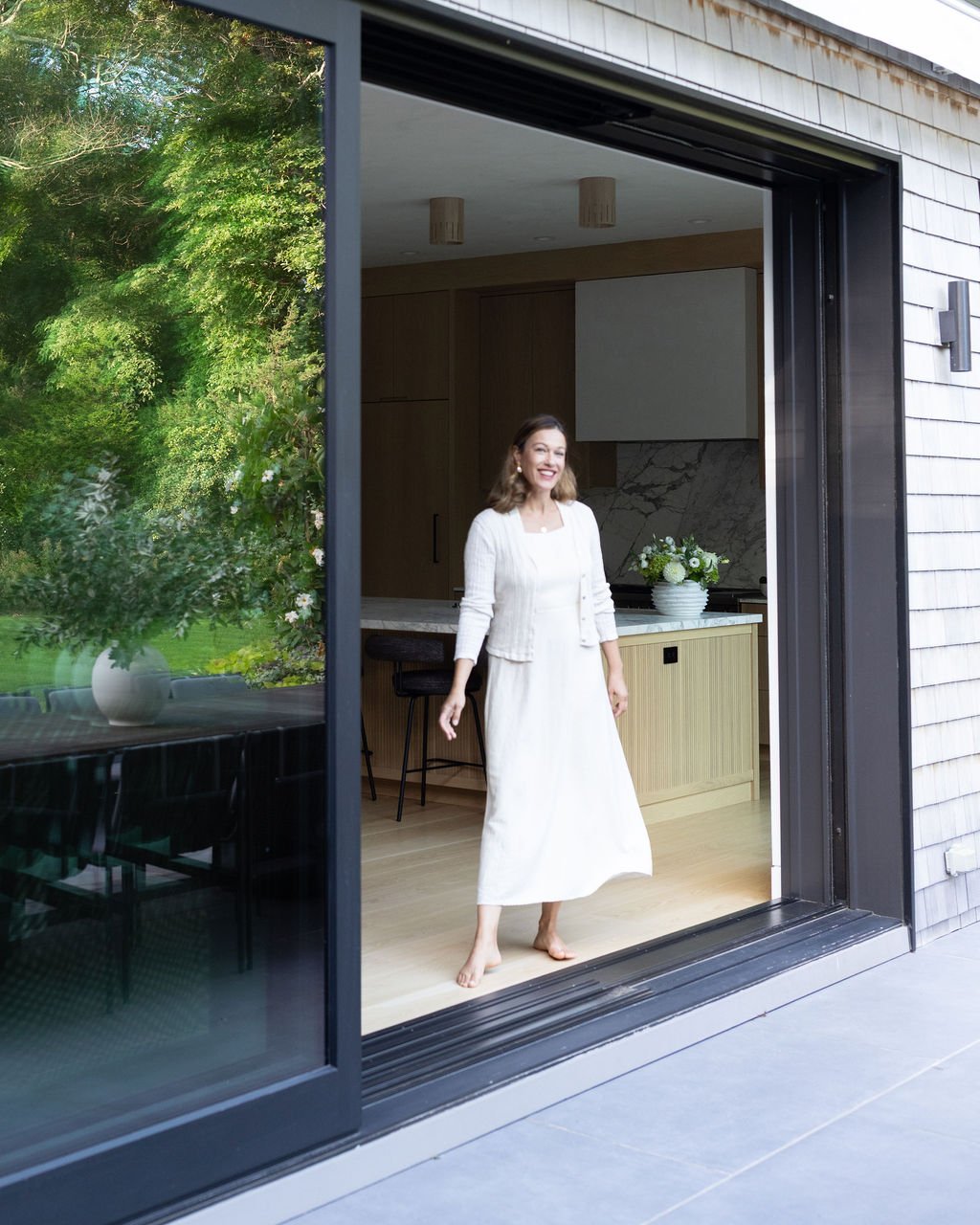 A woman standing barefoot, smiling inside a modern kitchen, seen through a sliding glass door with a reflection of green trees outside.