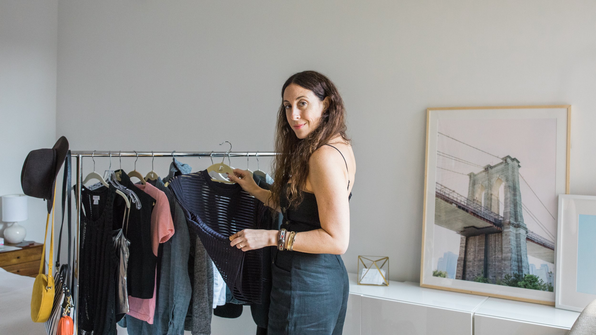 Woman with long brown hair shopping for clothes in a bedroom or dressing room, holding a striped black and gray dress, with a clothing rack filled with various garments nearby, a framed Brooklyn Bridge photo in the background.
