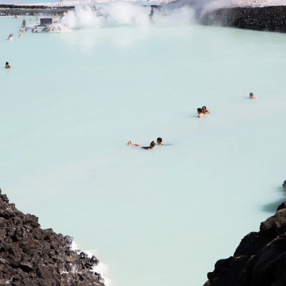 People swimming in a milky blue geothermal pool, with steam rising in the background and dark rocky edges surrounding the water.