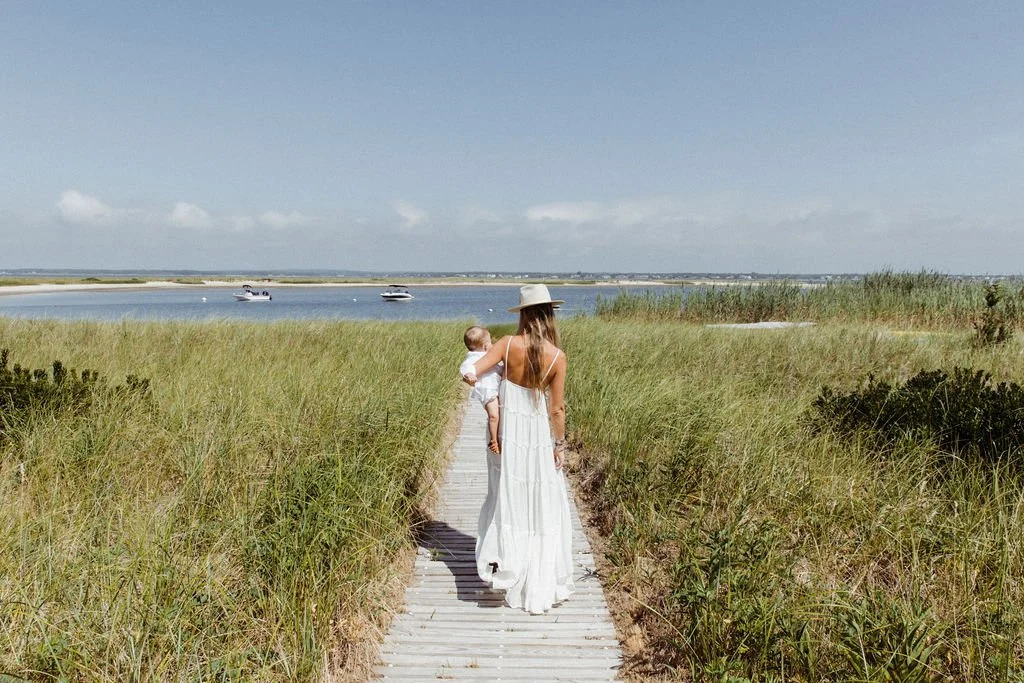 A woman in a white dress holding a child as they walk on a wooden path through grassy dunes towards a body of water with boats.