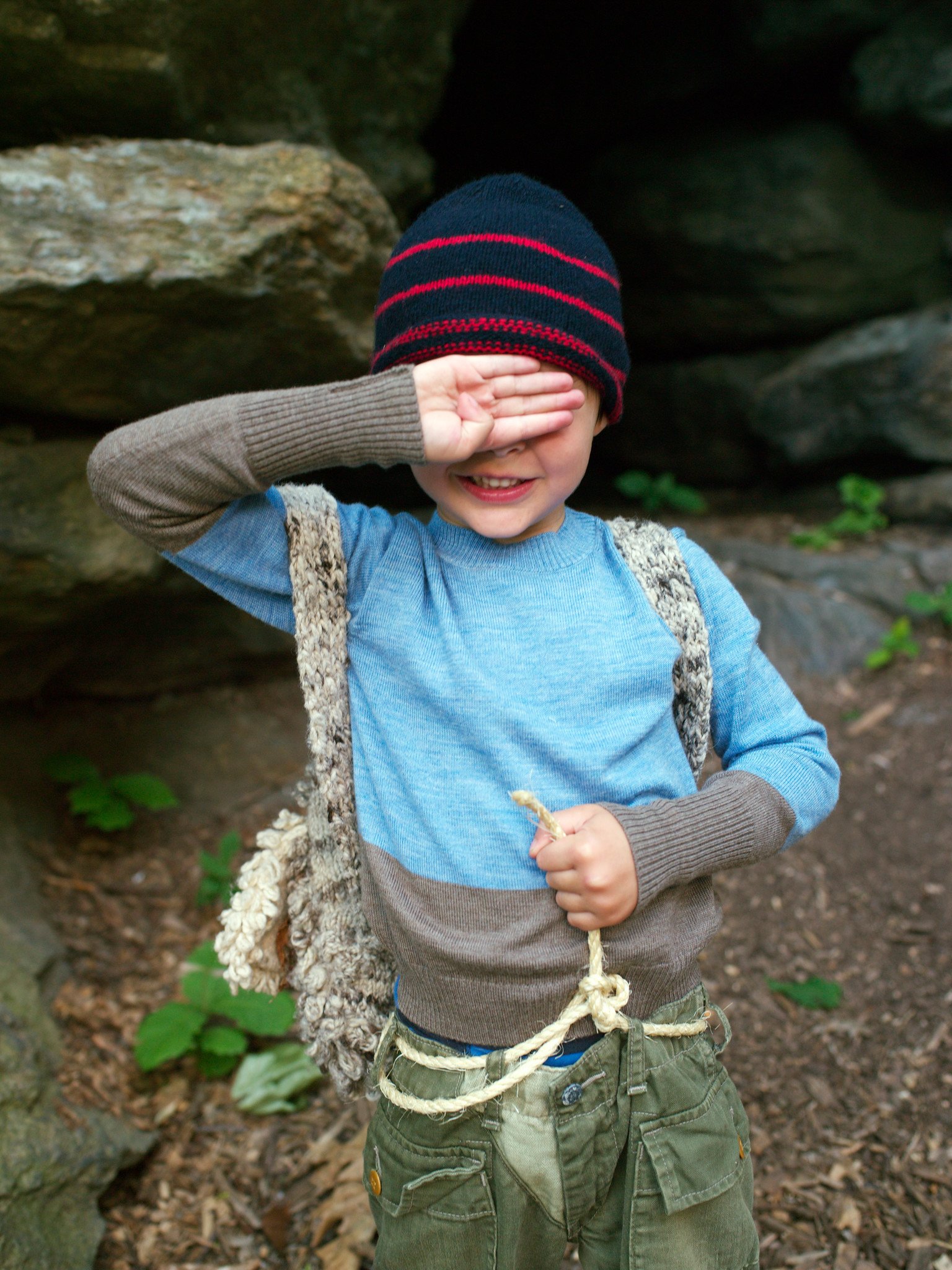 A young boy is outside in a wooded area, covering his eyes with his right hand and holding a rope in his left hand. He is wearing a striped beanie, a blue shirt with brown sleeves, and green cargo pants.