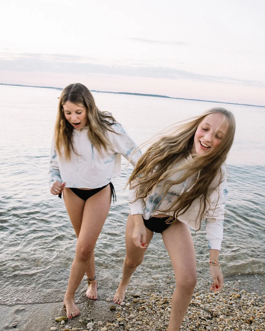 Two young women wearing hoodies and shorts standing at the edge of a lake, one smiling and the other looking surprised, as they prepare to step into the water.