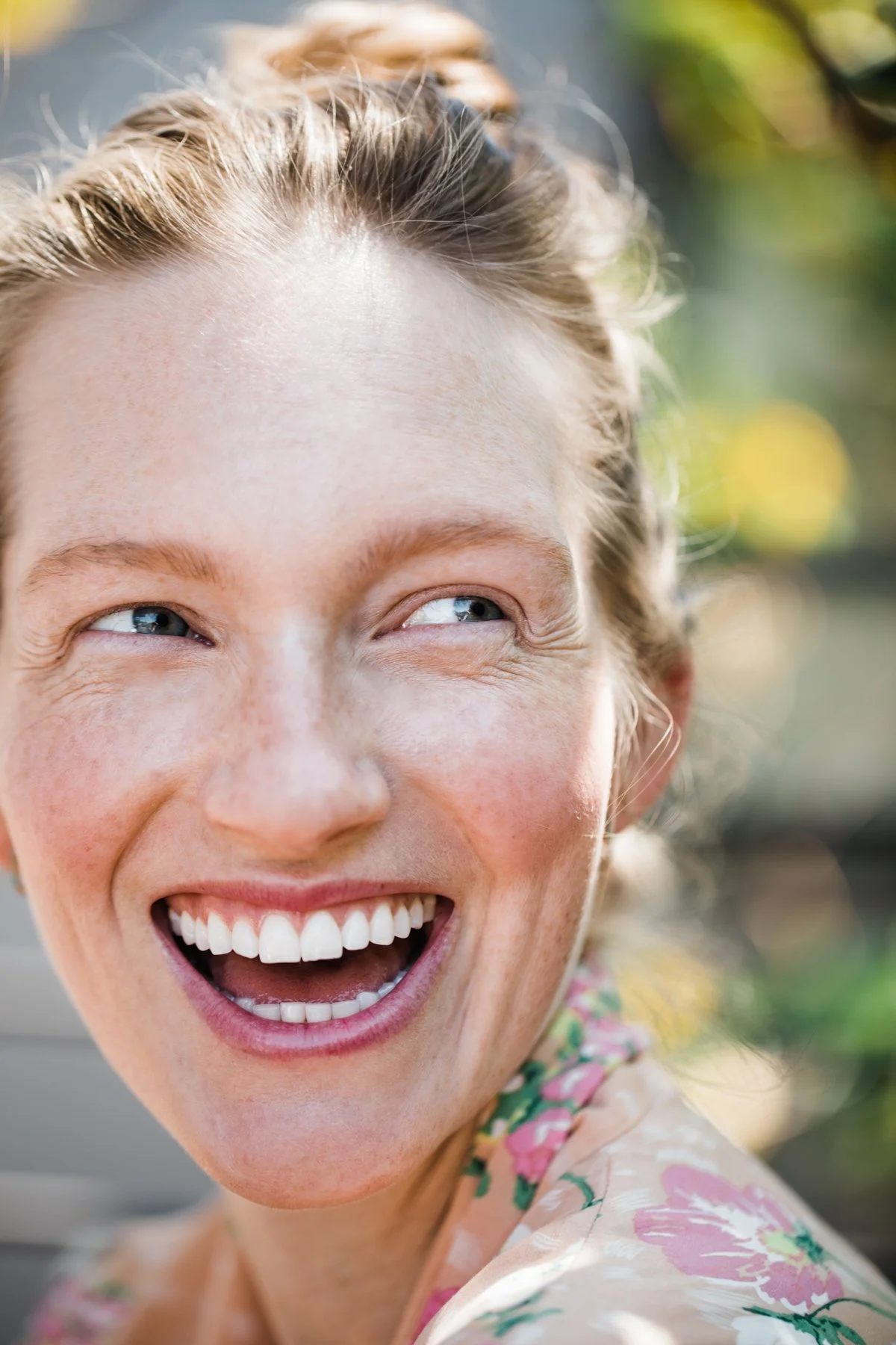 Close-up of a woman smiling and laughing outdoors, wearing a floral patterned top.
