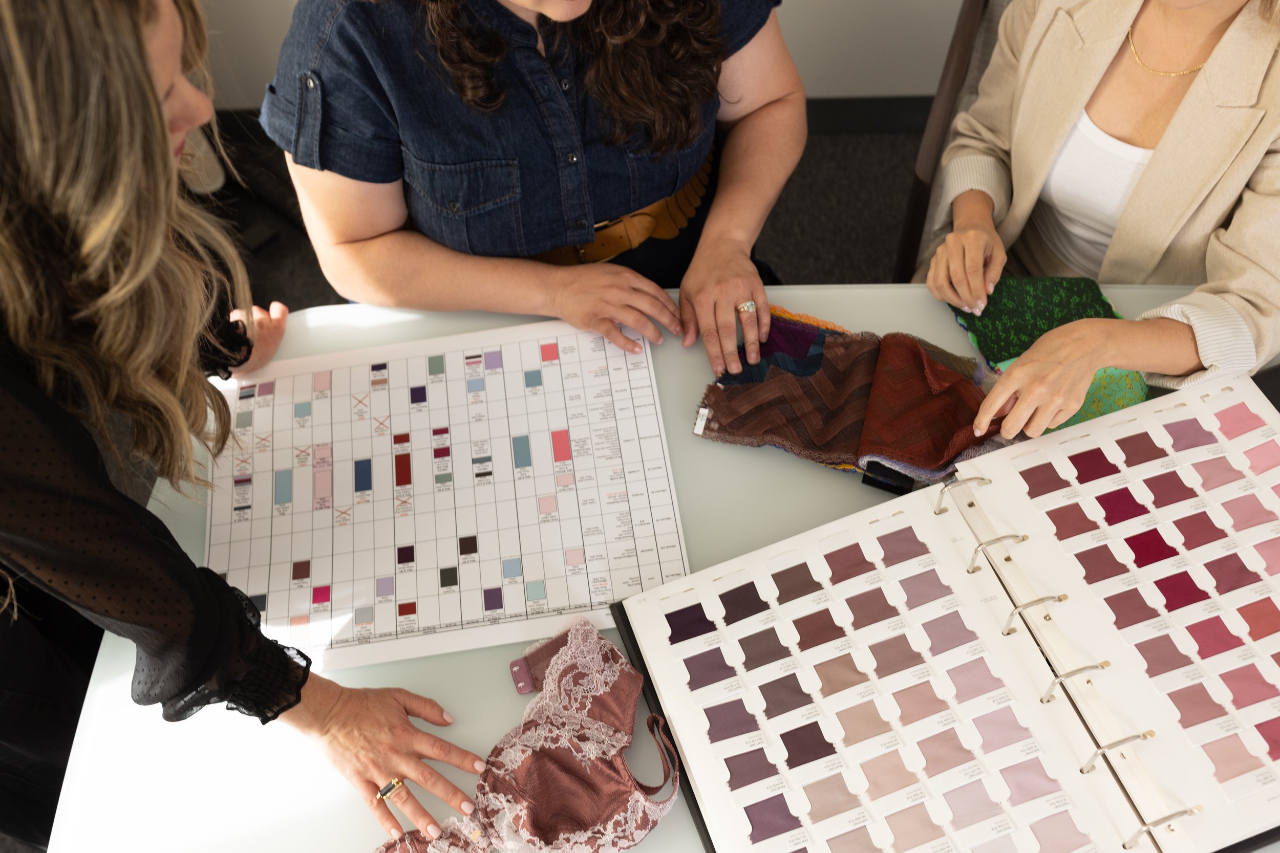 Three women are sitting at a table discussing fabric swatches and color charts, possibly planning a clothing or interior design project.