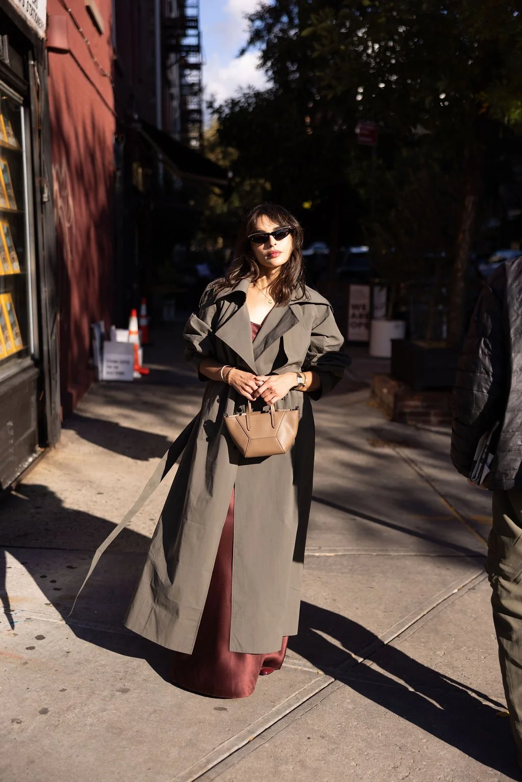 A woman wearing sunglasses and a brown trench coat standing on a city sidewalk in sunlight, holding a small tan handbag, with buildings and trees in the background.
