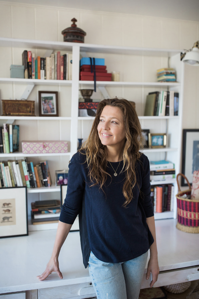 A woman with long wavy brown hair wearing a navy blue top and light blue jeans, standing in a room with white bookshelves filled with books and decorative items.
