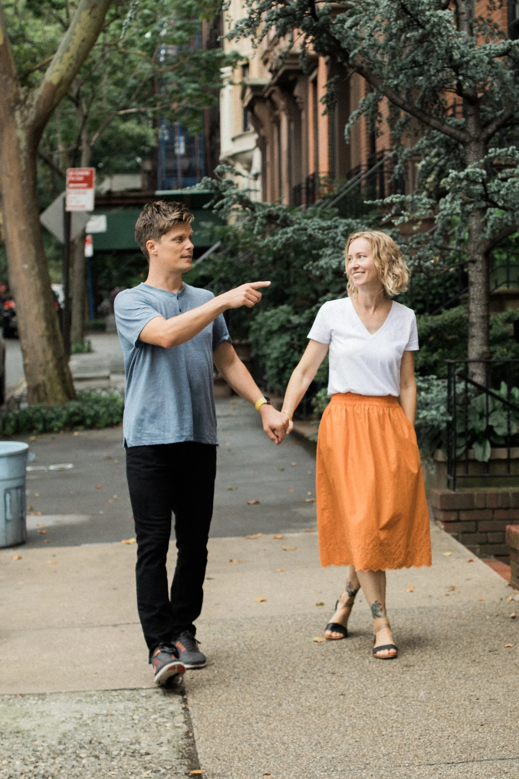 A young couple holding hands and walking on a city sidewalk park with trees. The man, wearing a blue T-shirt and black pants, is pointing at the woman. The woman, in an orange skirt and white top, is smiling and looking at the man.