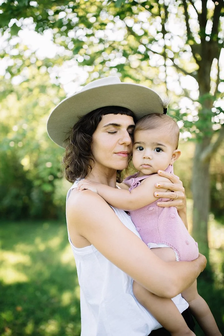 A woman wearing a beige hat holding a young girl in an outdoor park with green trees in the background.
