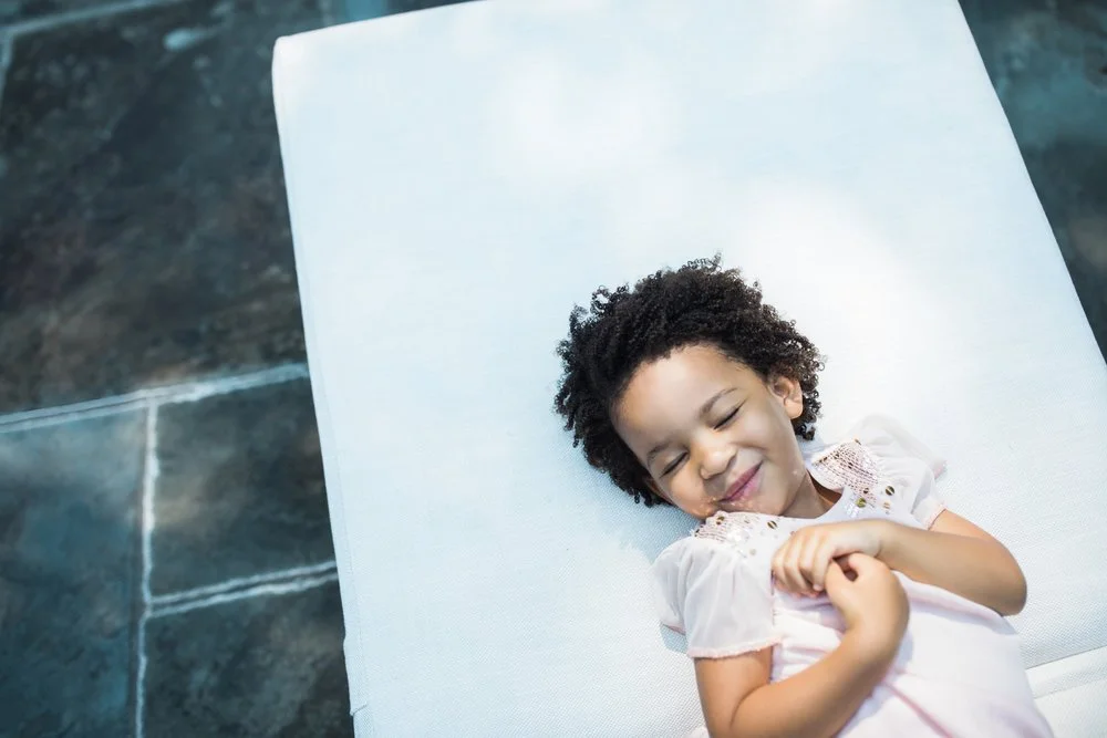 A young girl with curly hair smiling and lying on a white cushioned surface.