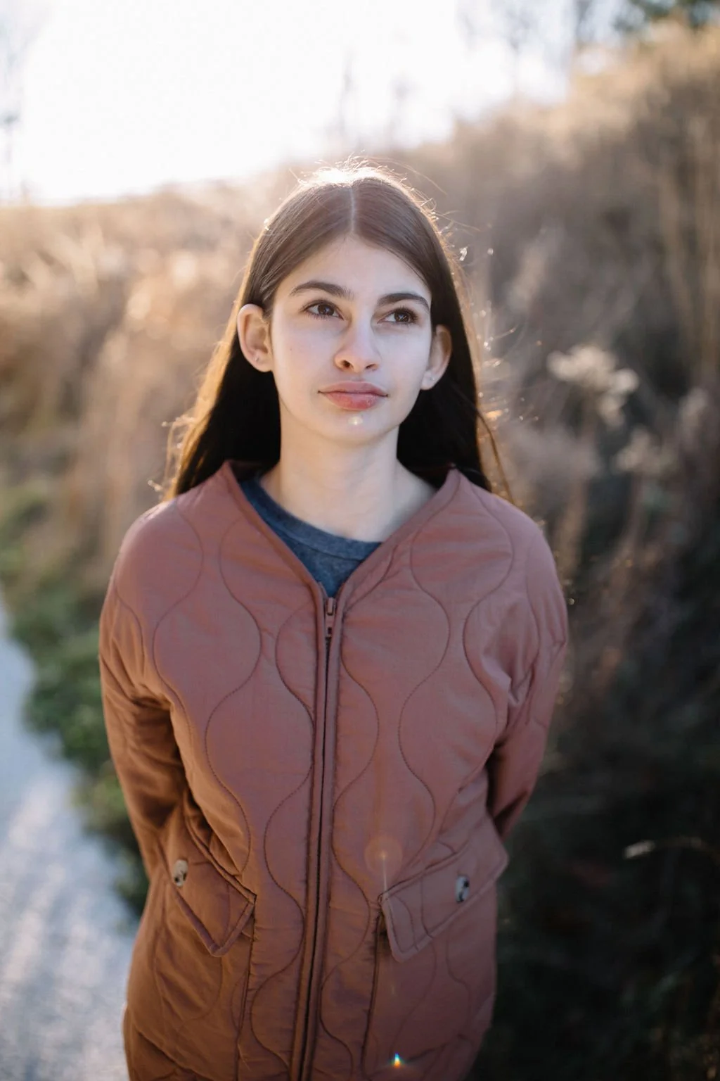 Young woman with long dark hair wearing a brown jacket standing outdoors in natural light with blurred background of trees and grass.