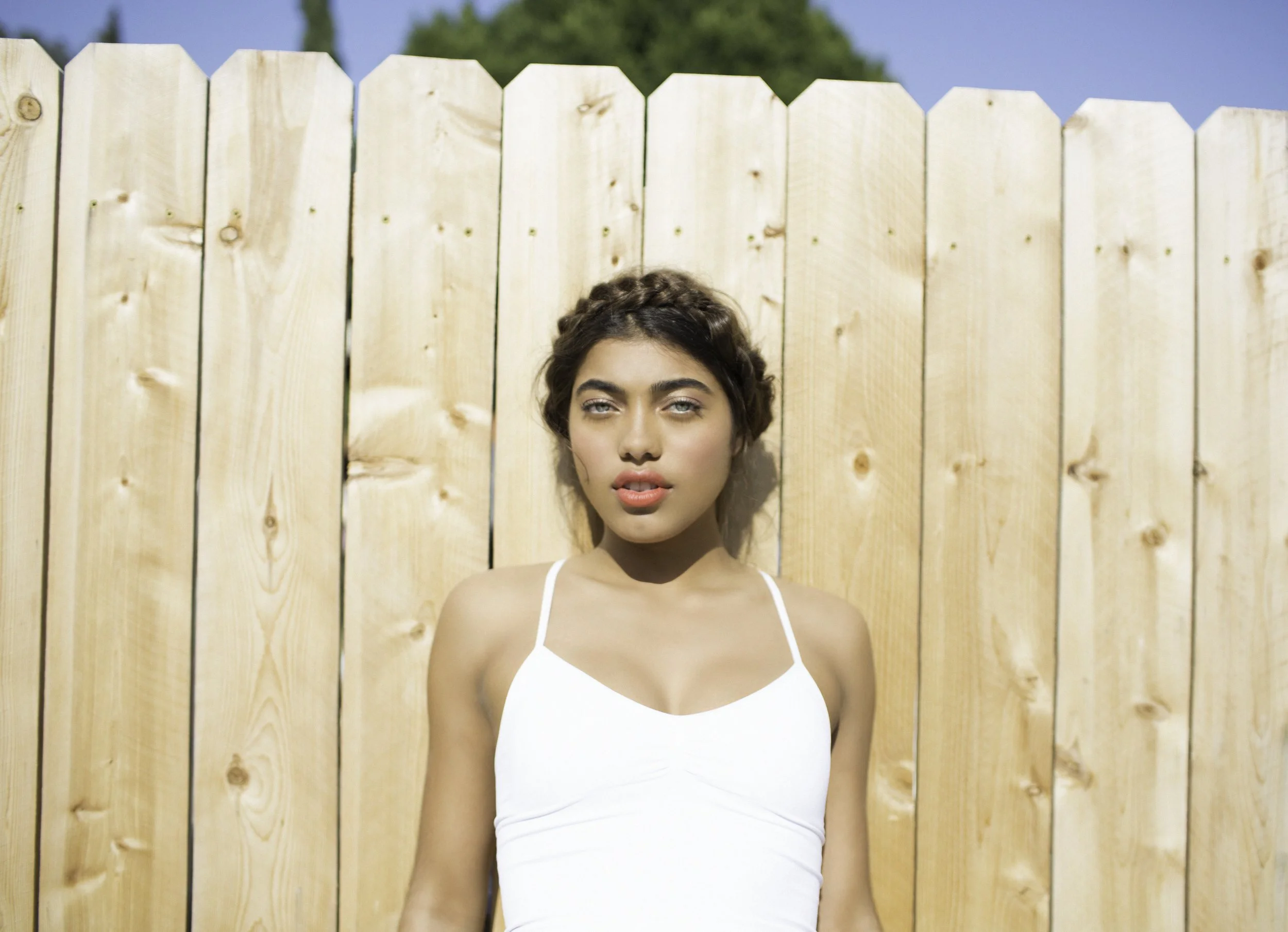 A woman in a white tank top standing outside in front of a wooden fence with blue sky and green trees in the background.