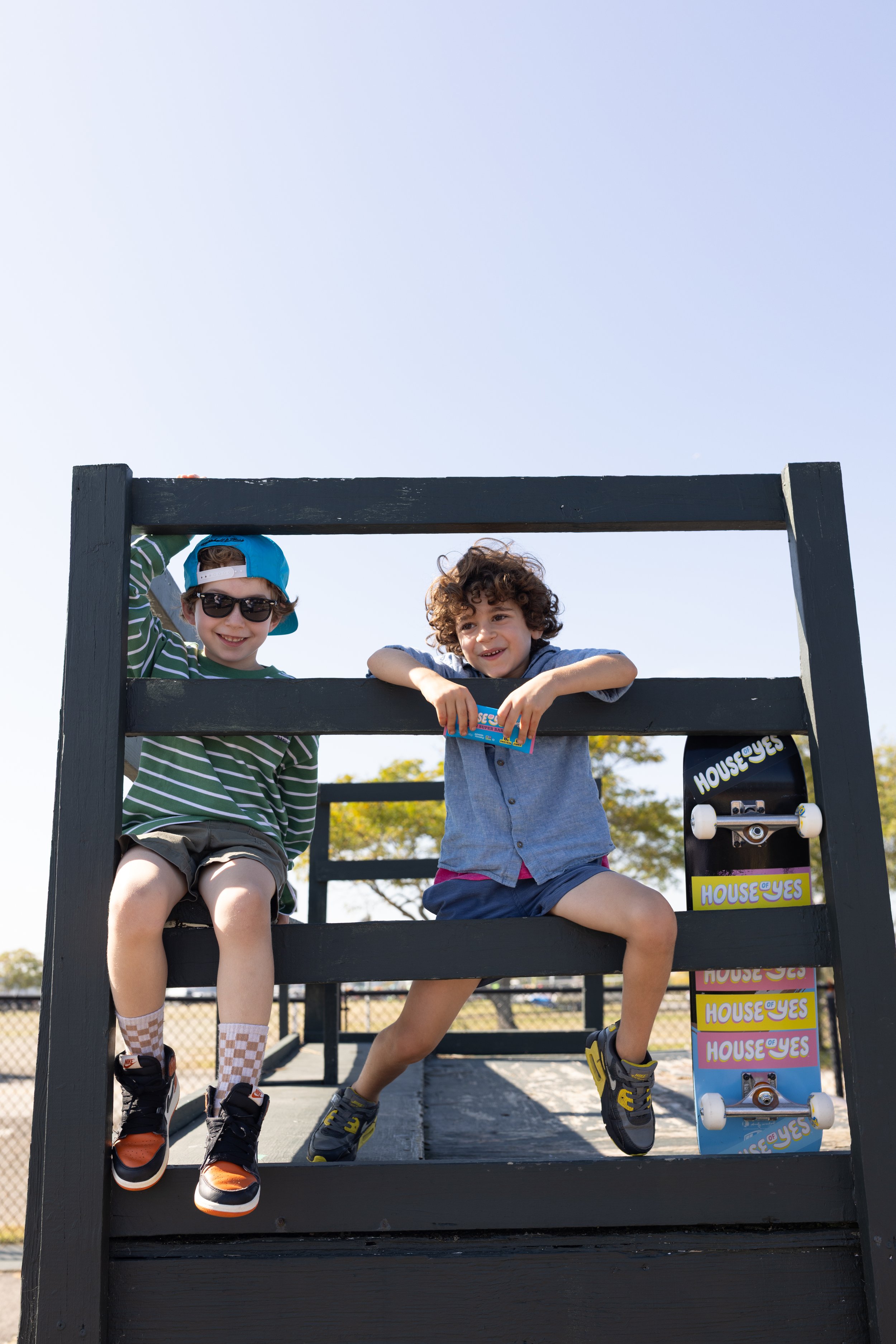 Two young boys sitting on a playground structure under a clear sky, one wearing sunglasses, a blue cap, and striped shirt, the other with curly hair and a gray shirt, holding a small toy.