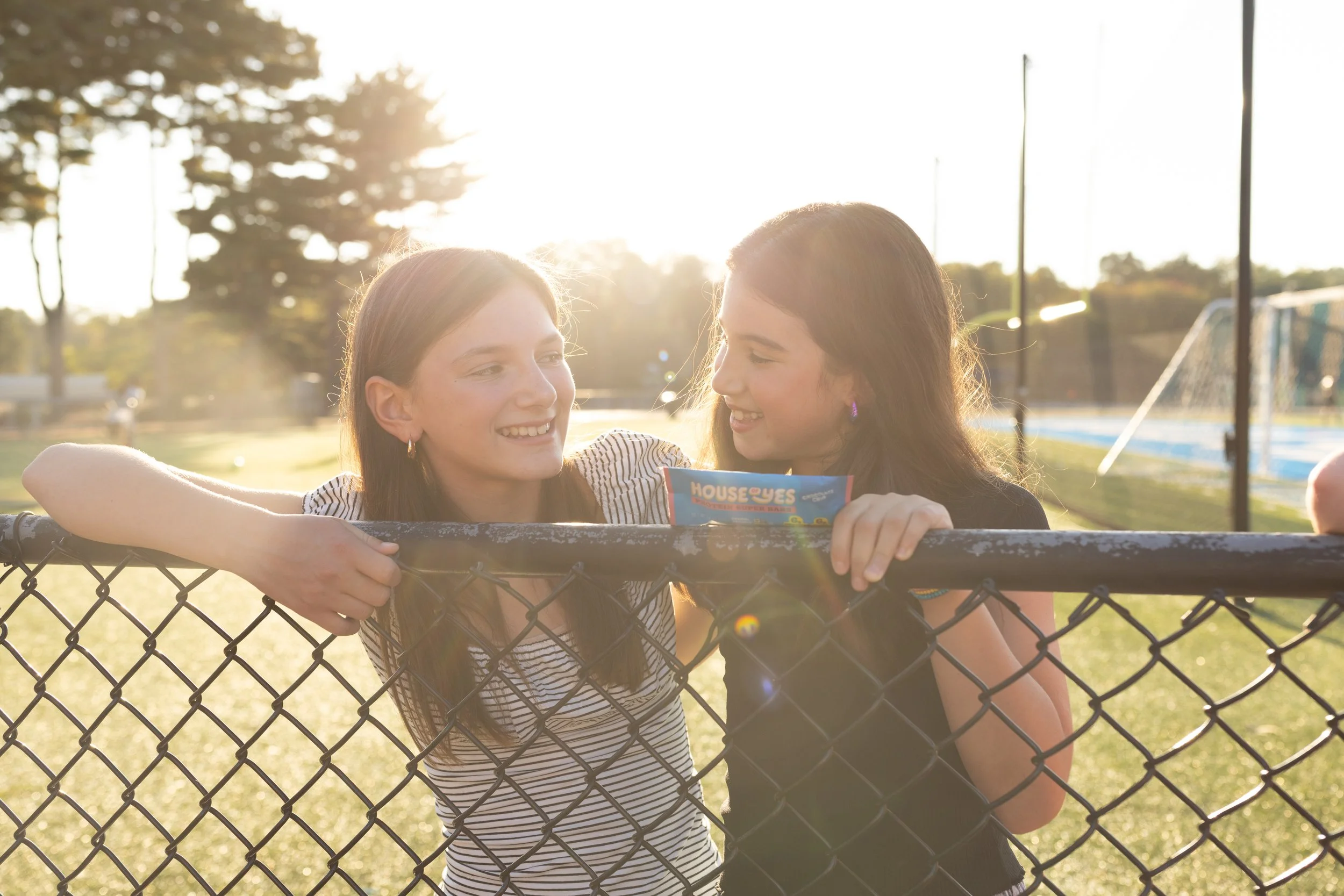 Two smiling girls leaning on a chain-link fence at a park during sunset, sharing a moment of happiness.
