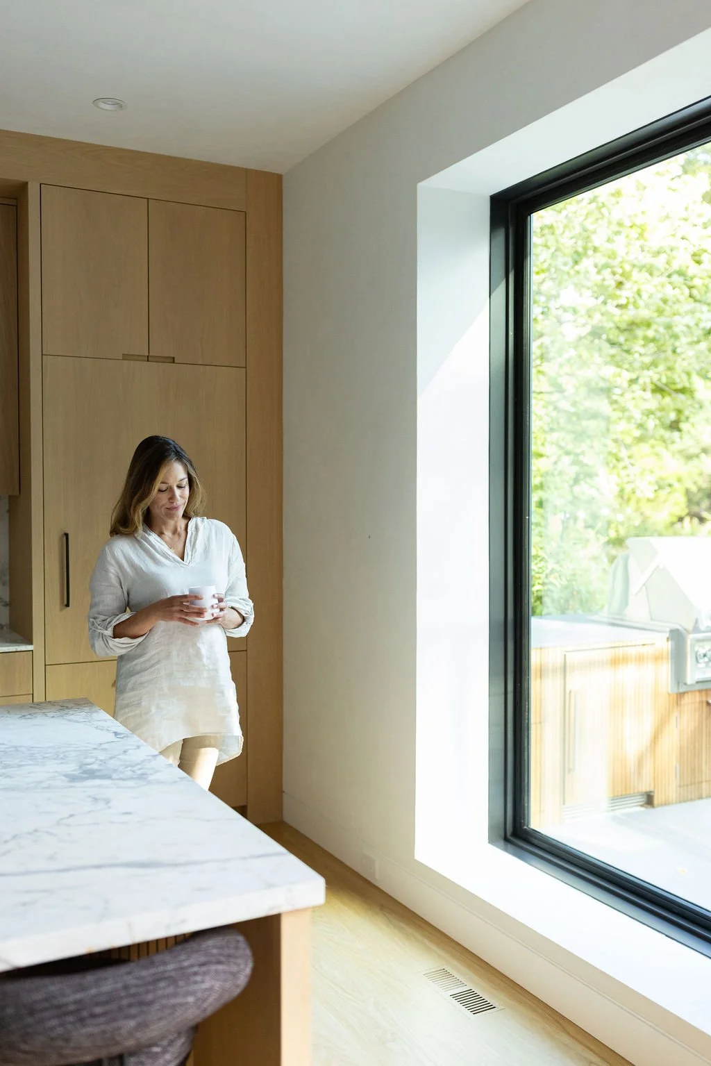 A woman in a white blouse holding a cup in a modern kitchen with light wood cabinets, a marble countertop, and large glass sliding door overlooking a green backyard.