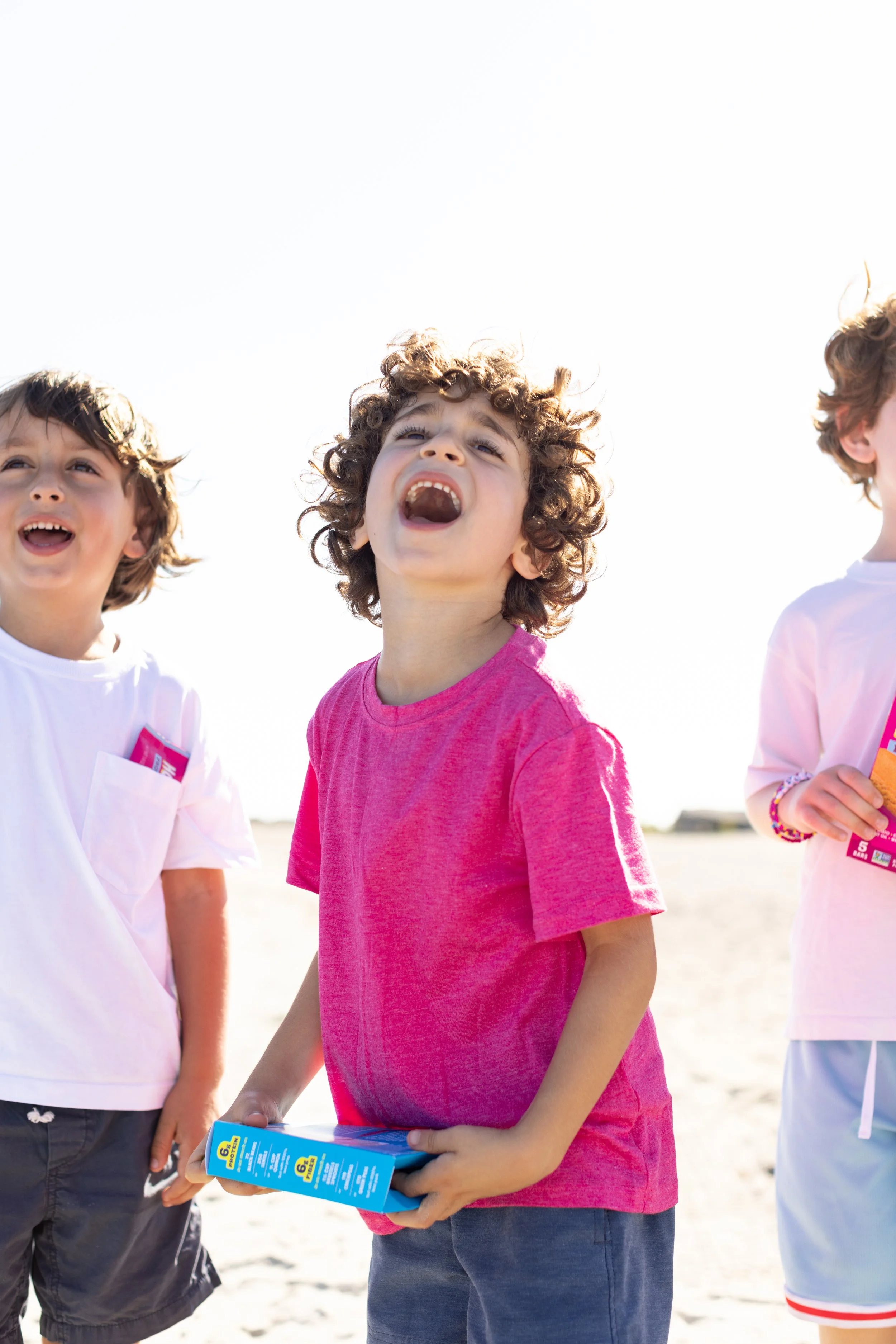 Three children standing on a beach, one girl in the center with curly hair wearing a pink shirt holding a box, with two boys next to her, one in a white shirt and the other in a pale pink shirt, partly visible.