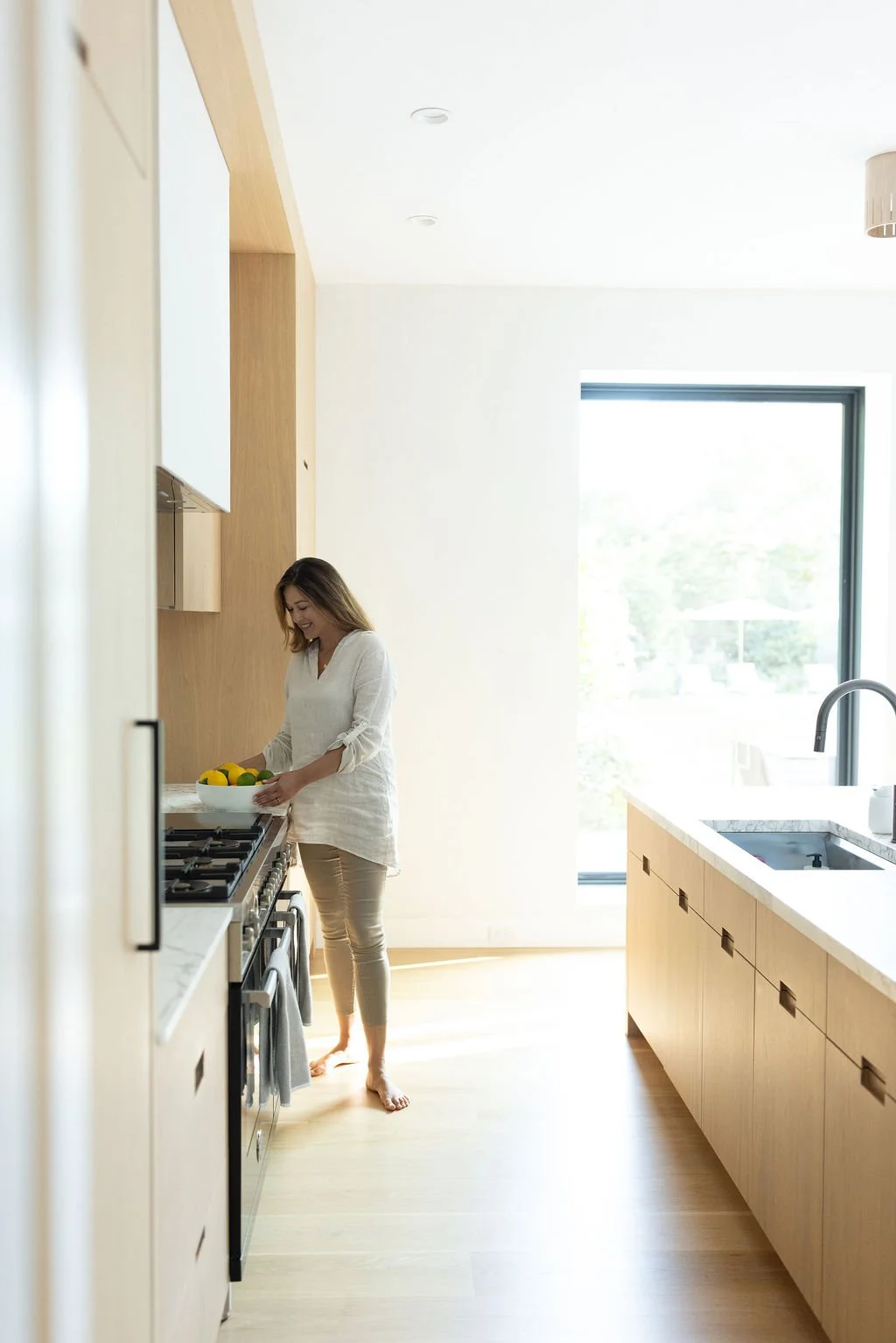 A woman standing barefoot in a bright, modern kitchen, holding a bowl of lemons and limes near the stove.