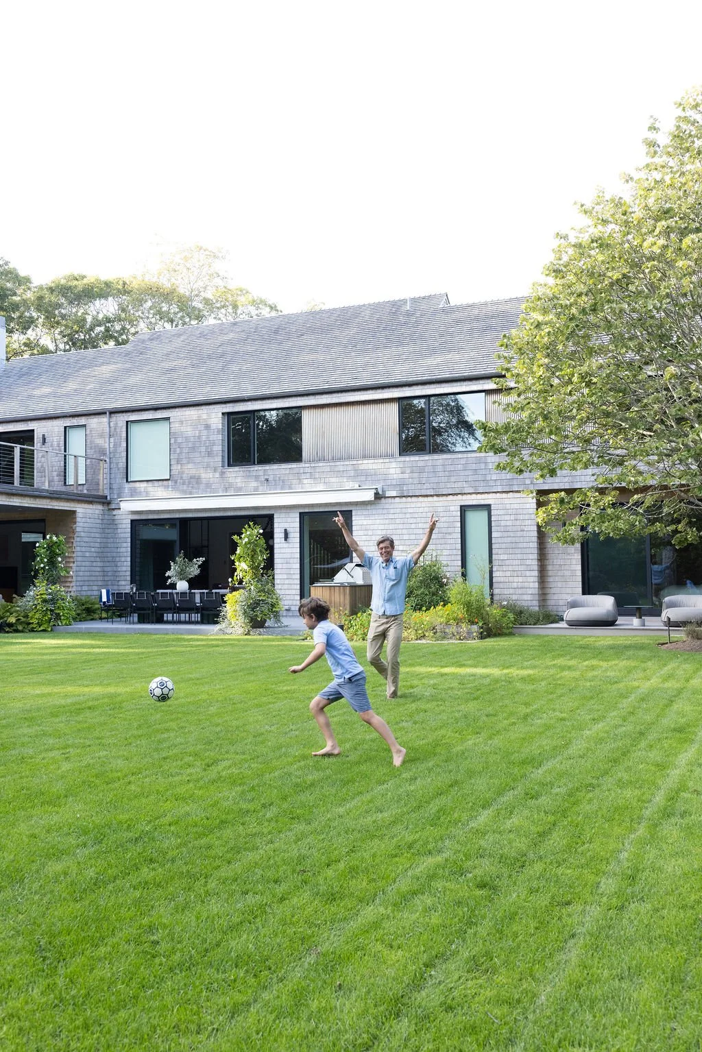A man and a boy playing with a soccer ball on a well-maintained green lawn in front of a modern house with large windows and a patio area, during daytime.