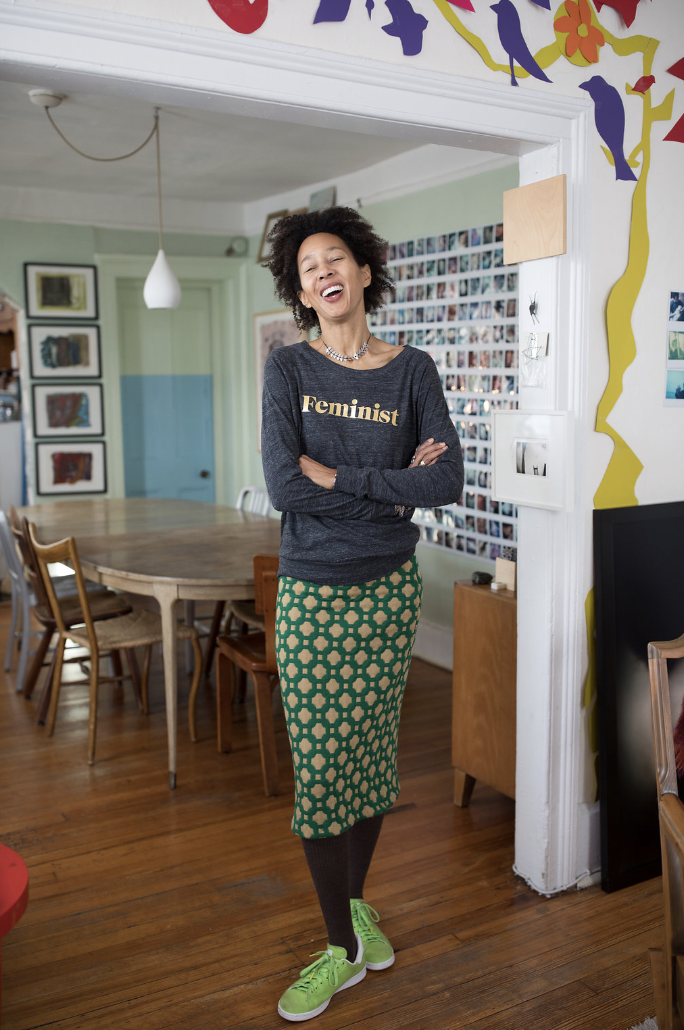Woman standing in a decorated indoor space, smiling with arms crossed, wearing a shirt with 'Feminist' written on it, patterned green skirt, and bright green sneakers.