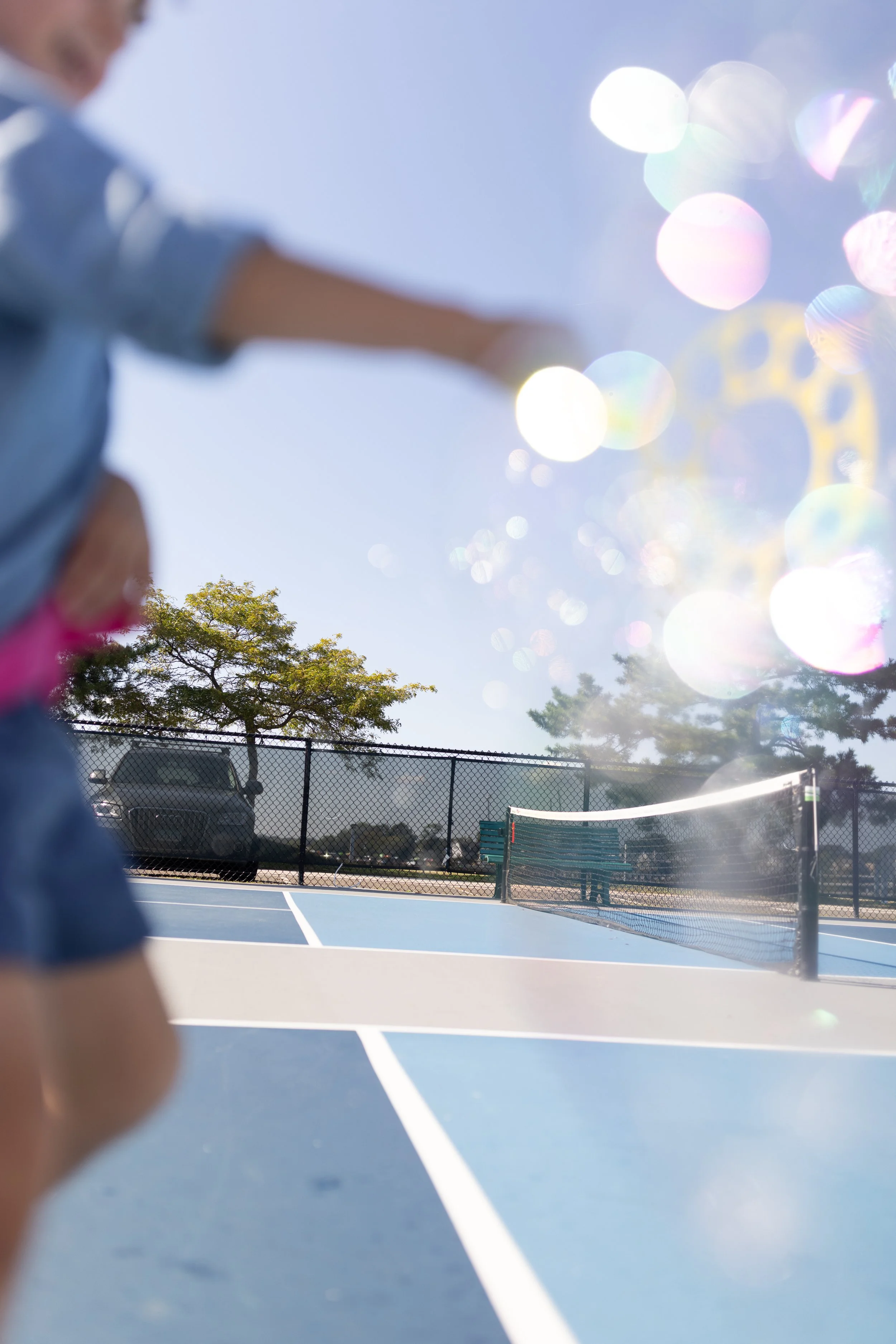 A blurry photo of a person on a tennis court with a background of trees, a parked car, and a fence, with sunlight creating lens flare.