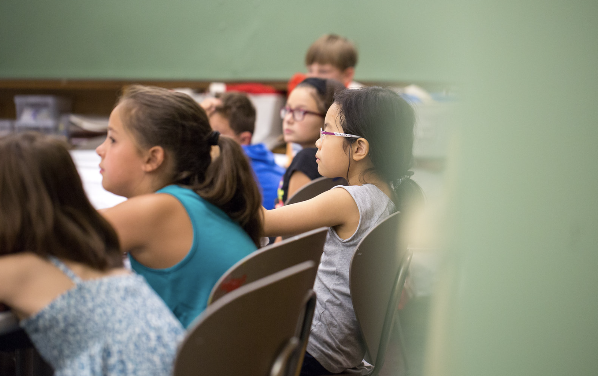 Children sitting in a classroom, attentively listening, with a green chalkboard in the background.