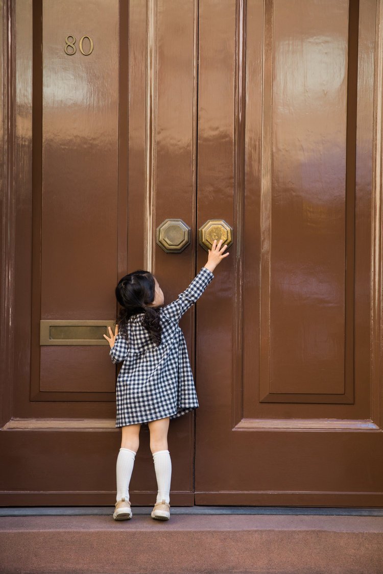 A young girl in a black and white checkered dress and knee-high white socks reaching for the brass doorknob of a large brown wooden door.
