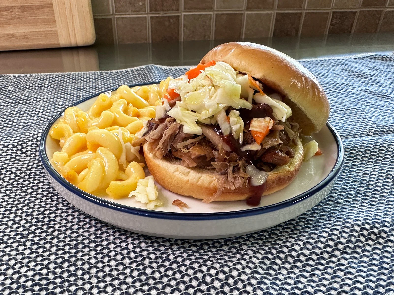 A plate with a pulled pork sandwich topped with bbq sauce and coleslaw with a side of macaroni and cheese