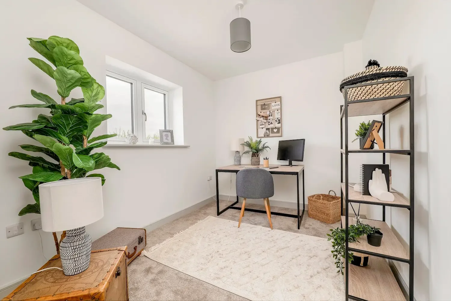 A minimalist home office with a large green potted plant, a wooden laptop stand, a small desk with a computer monitor, a gray chair, a black metal shelf with decorative items, a white rug, and a window providing natural light.