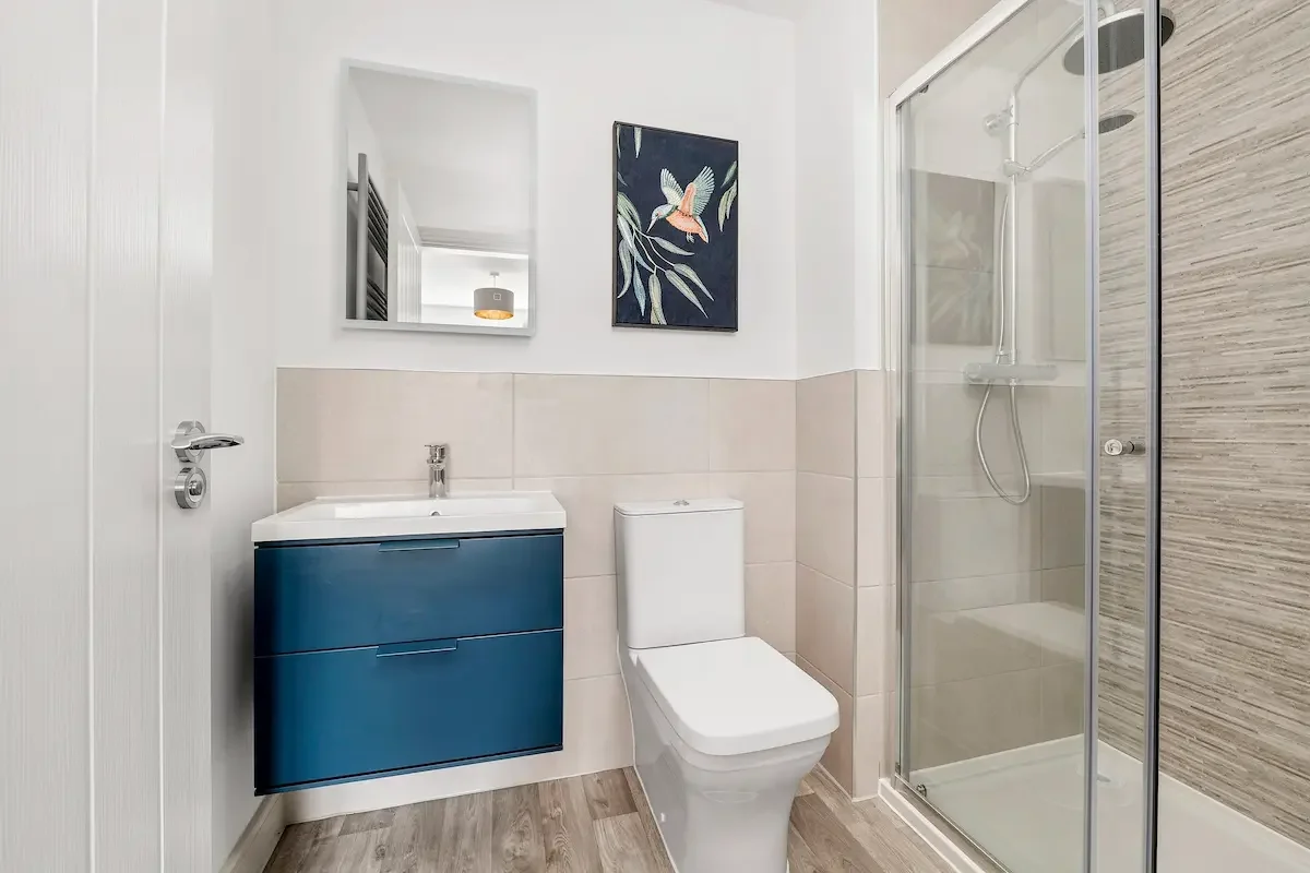 Modern bathroom with a blue vanity, white sink, white toilet, a framed artwork of a bird, a mirror, and a glass shower enclosure with wood-textured tiles inside.