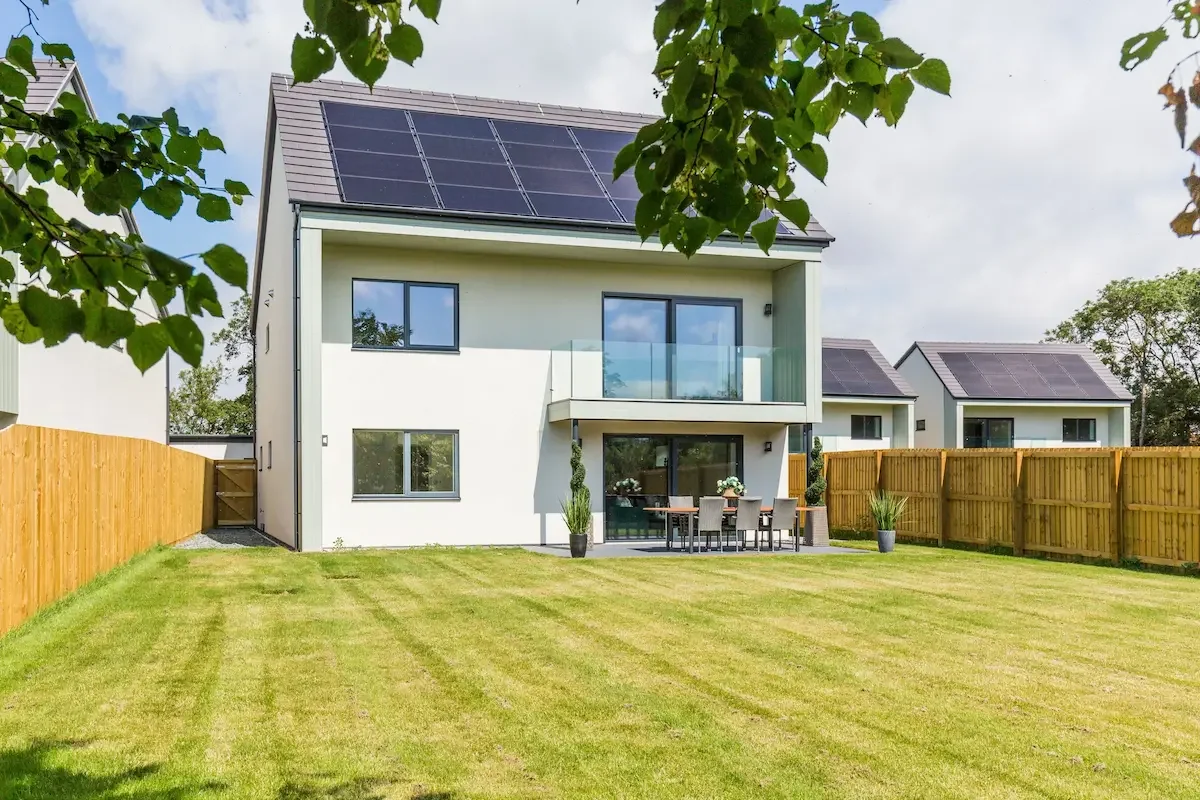 A modern white house with solar panels on the roof, a balcony, and outdoor seating area, situated on a massive green garden