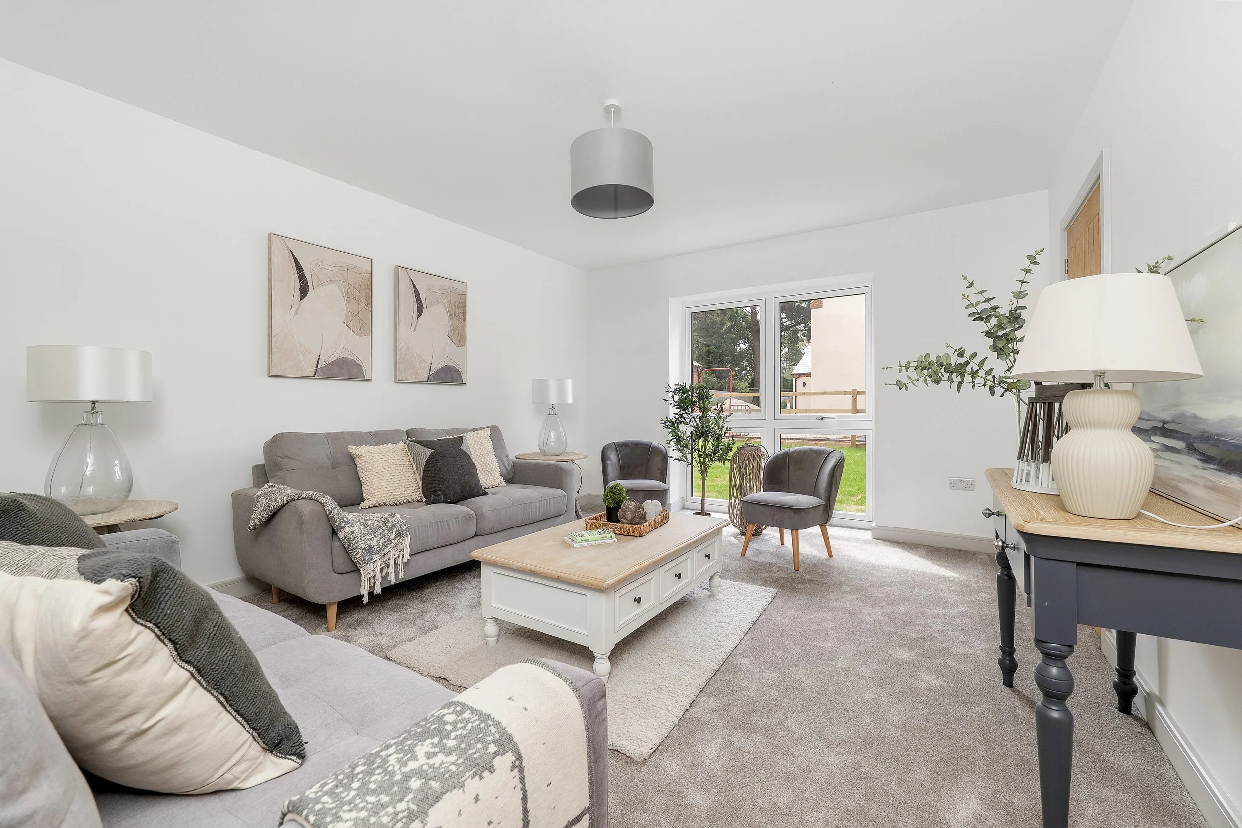 A cozy, neutral-toned lounge with grey sofas, plush carpeting, and a white wooden coffee table. Large windows provide a view of a green lawn and a wooden fence outside.