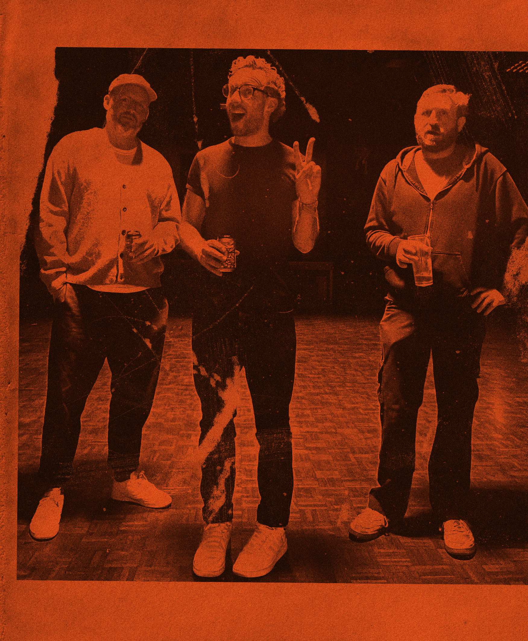 Three men standing together in a dimly lit indoor space, each holding a drink, with the man in the center making a peace sign gesture. The photo has a reddish-orange filter applied.