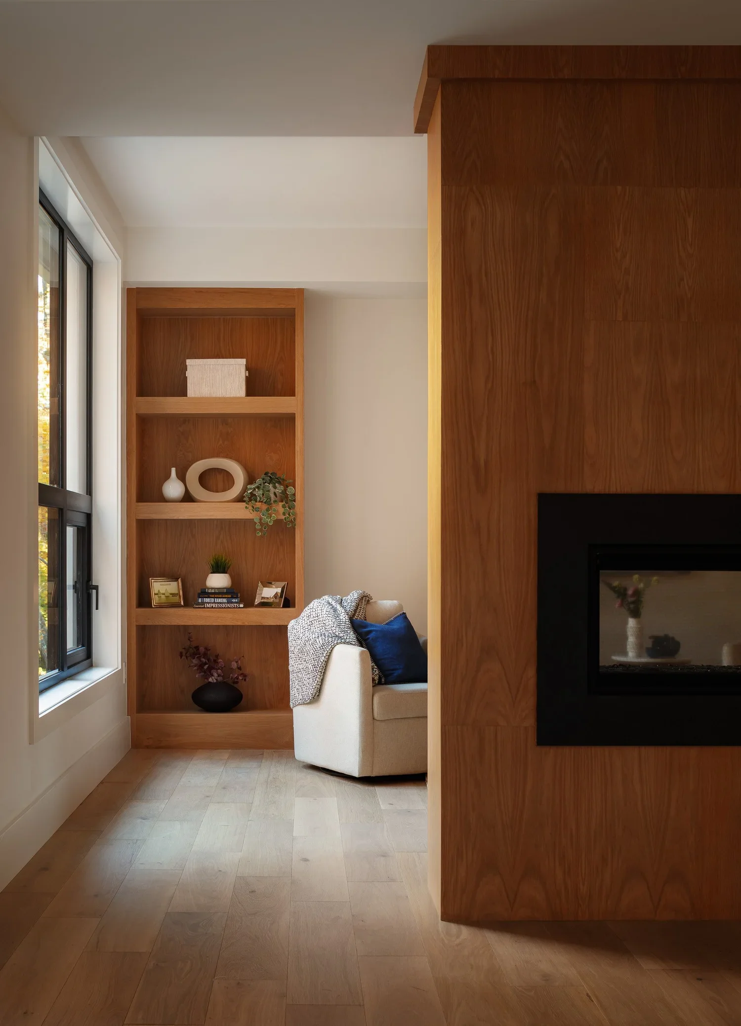 A cozy living room corner featuring a beige sofa with pillows, a white curtain, a wooden bookshelf with decorative items, and a fireplace framed with black. The floor is light wood, and the room is illuminated by natural light from a large window.
