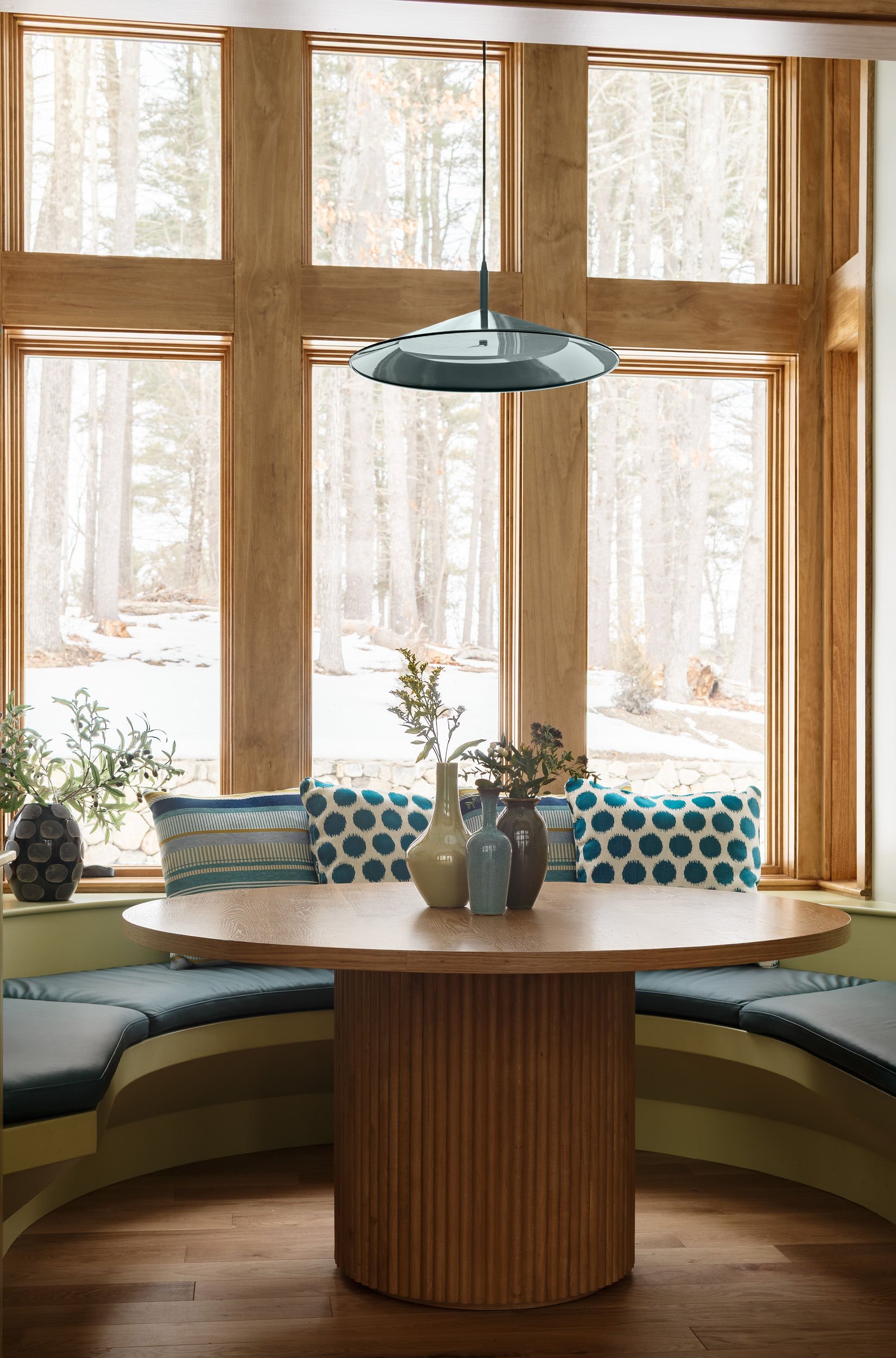 A cozy corner inside a wooden house with large window view of snow-covered trees, a round wooden table with decorative vases, cushioned seating, and cushions with polka dot and striped patterns.