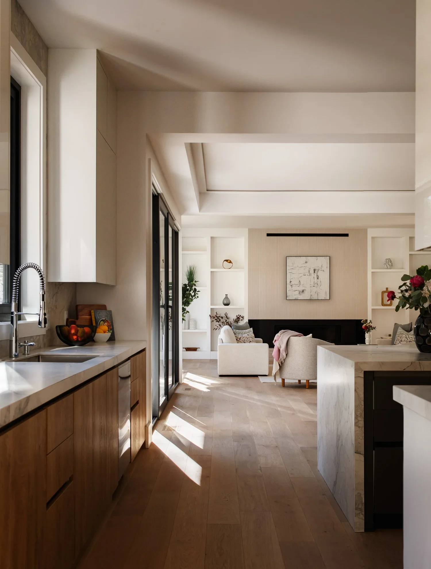 Modern open-concept kitchen and living room with natural light, white walls, wooden flooring, kitchen island, beige sofa, built-in shelves, and decorative items.