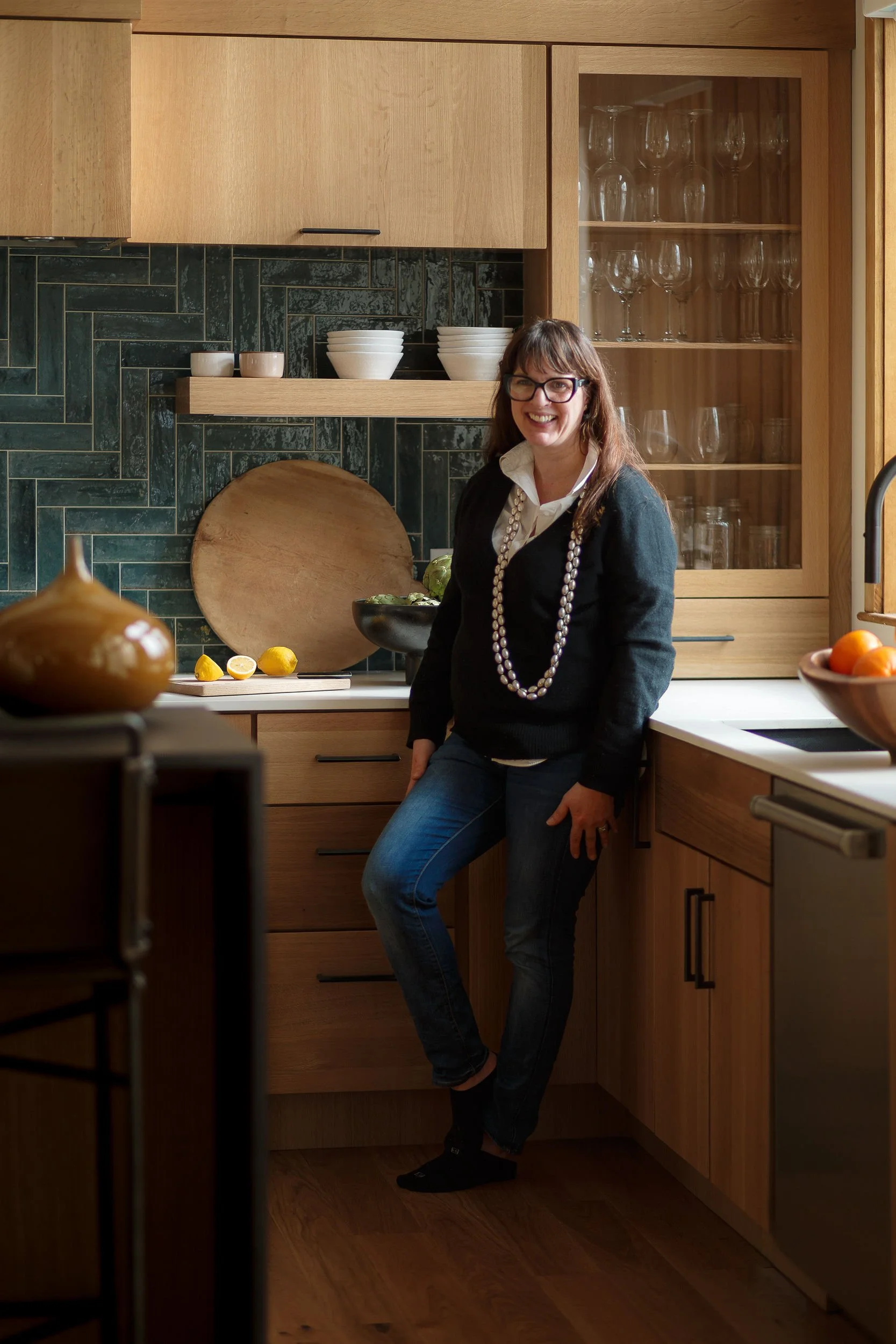 A woman with glasses and a pearl necklace smiling in a modern kitchen with wooden cabinets, green tile backsplash, and a black countertop. There are bowls, lemons, and glassware visible in the background.