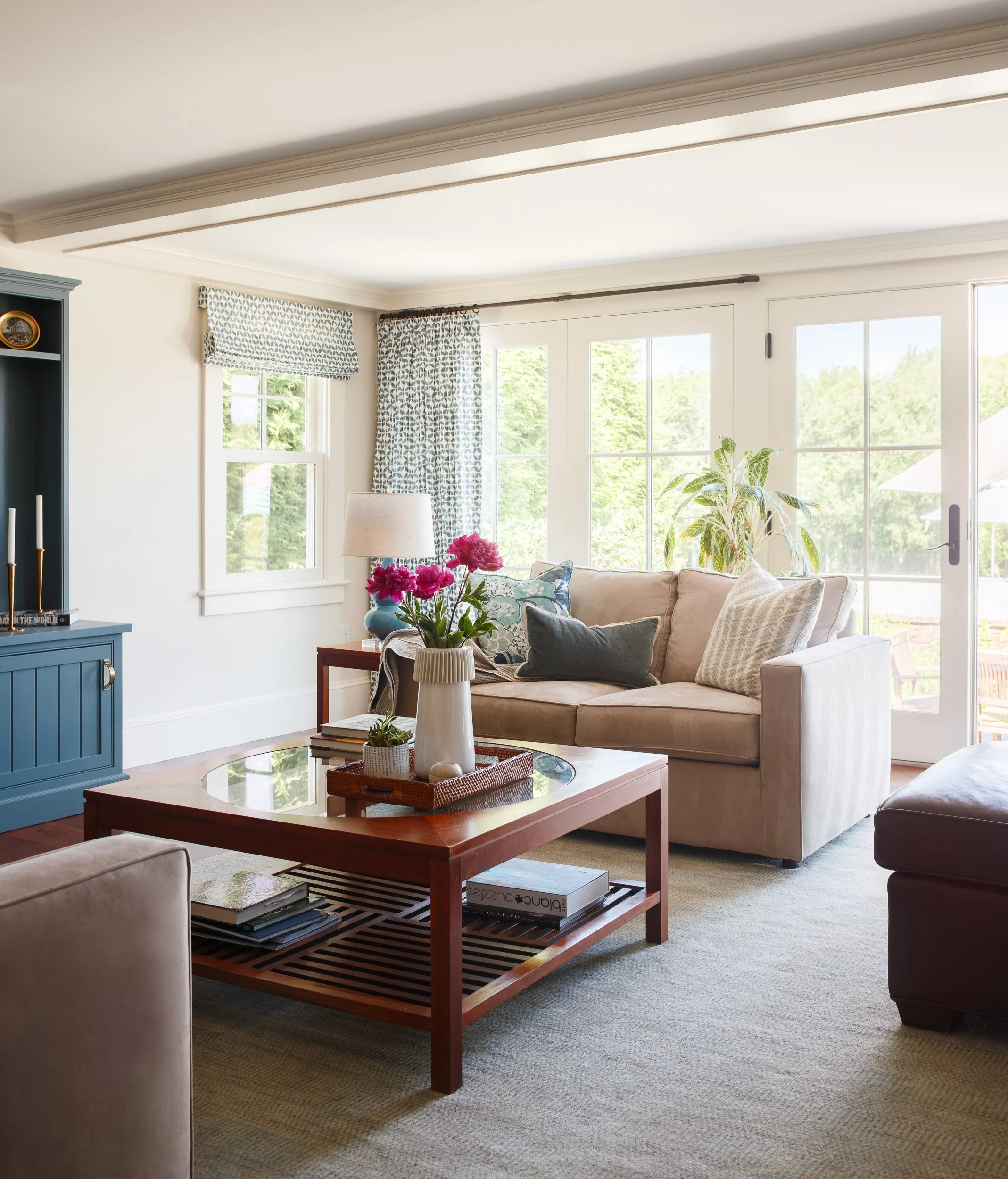 A cozy living room with large windows and beige furniture, including a sofa with cushions, a coffee table with flowers, and a blue cabinet, decorated with patterned curtains and a bookshelf.