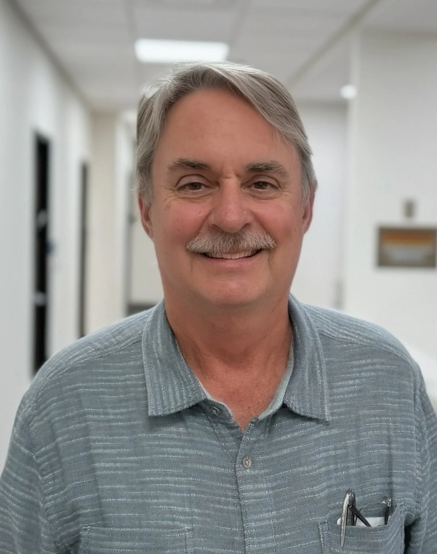 A smiling middle-aged man with gray hair and a mustache wearing a gray collared shirt standing in a hallway.