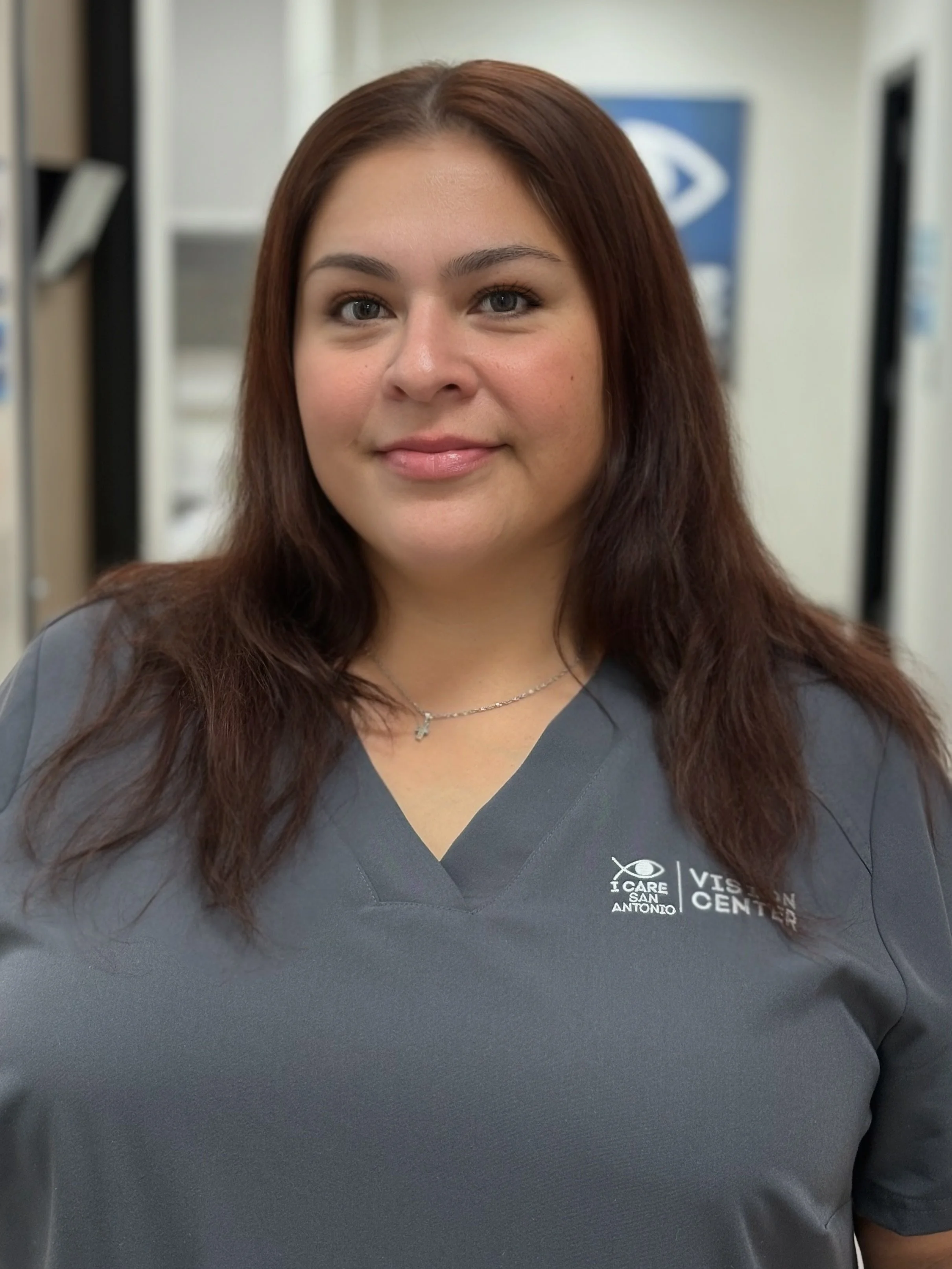 A woman with long dark hair, wearing a gray healthcare uniform with a logo, smiling in a medical or office setting.