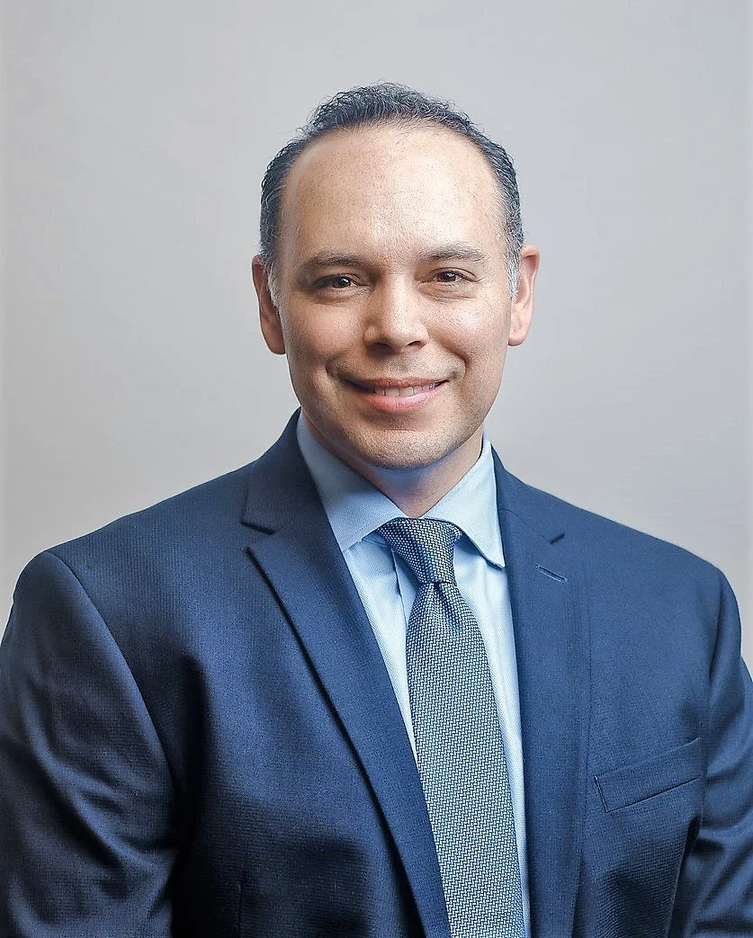 Professional headshot of a man in a navy suit, light blue shirt, and patterned tie, smiling against a neutral background.