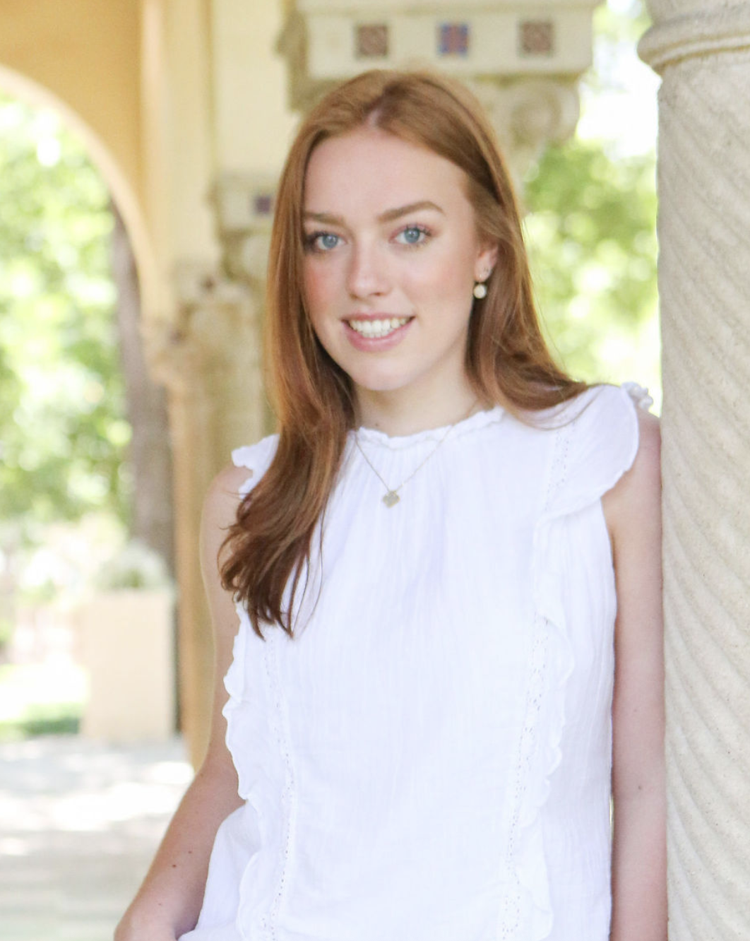 A young woman with long red hair and blue eyes smiling, wearing a white sleeveless top, standing outdoors next to a textured stone column, with greenery and architectural elements in the background.