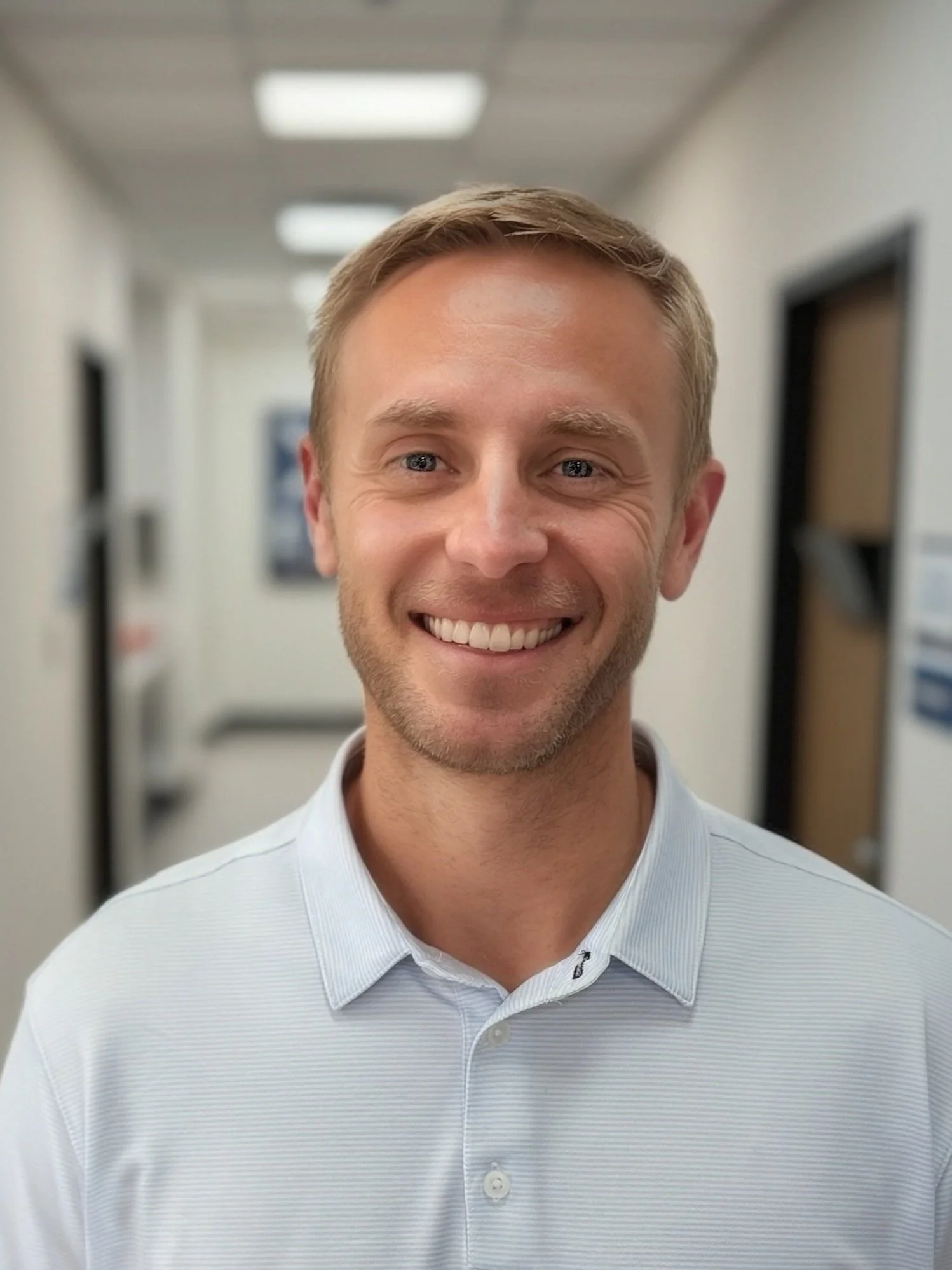 A smiling man with light brown hair and a beard, wearing a white collared shirt, standing in a corridor with blurred background.