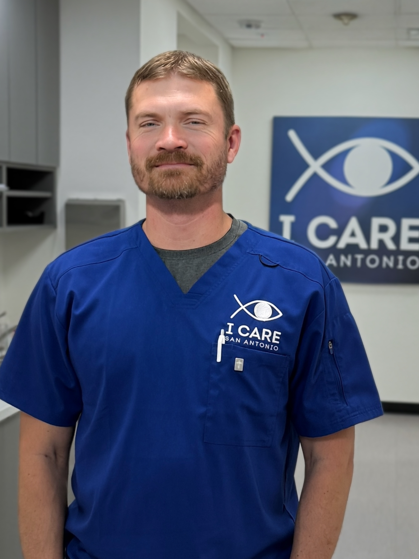 A man with short brown hair and a beard, wearing a light blue striped button-up shirt, standing outdoors in front of green leafy bushes.