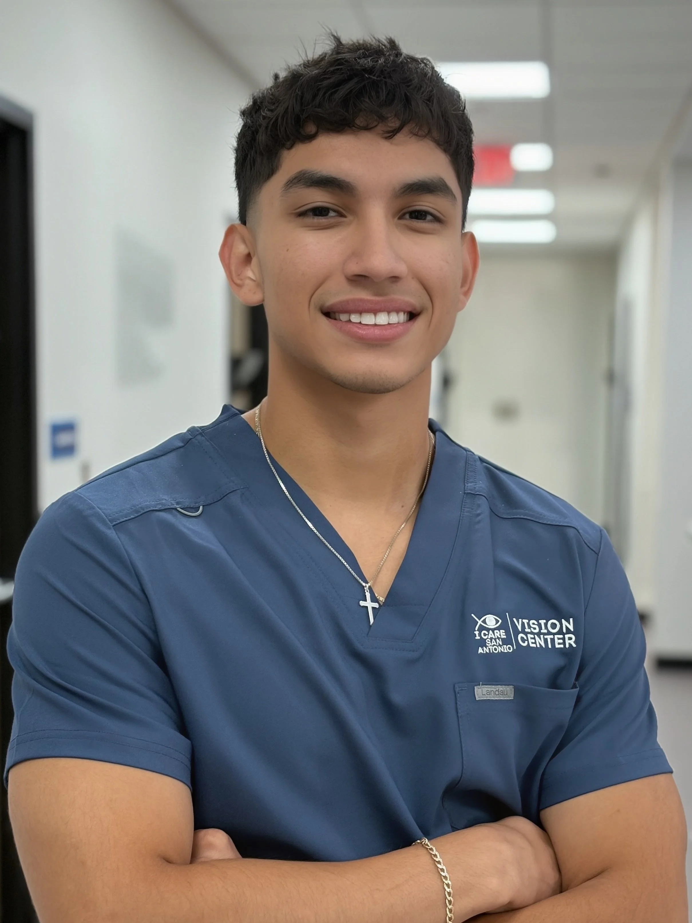 Young man in blue scrubs with cross necklace and bracelet, standing in a medical facility, smiling with arms crossed.