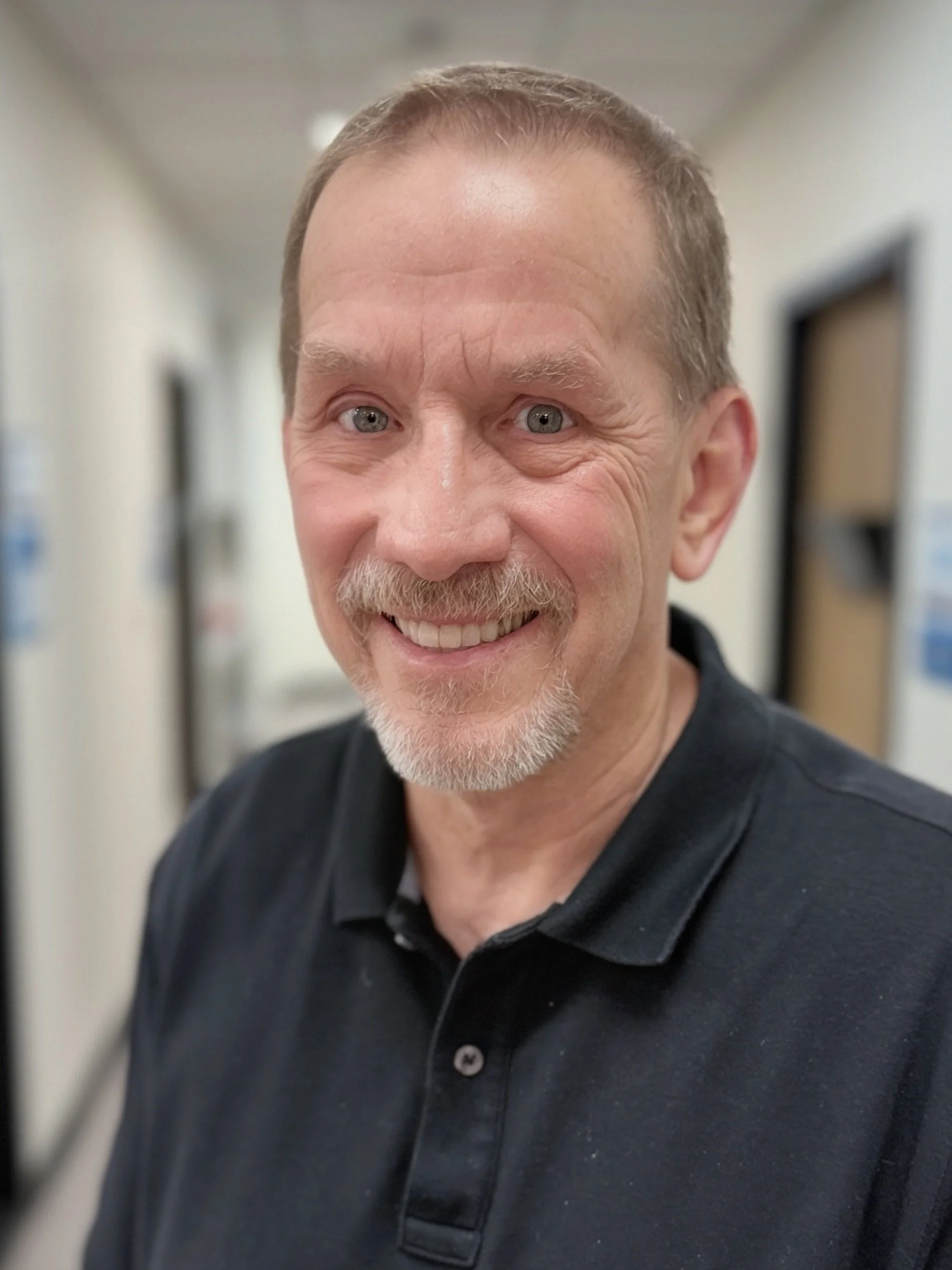 A smiling middle-aged man with short light brown hair, a beard, and blue eyes wearing a black polo shirt, standing in a hallway with doors on either side.