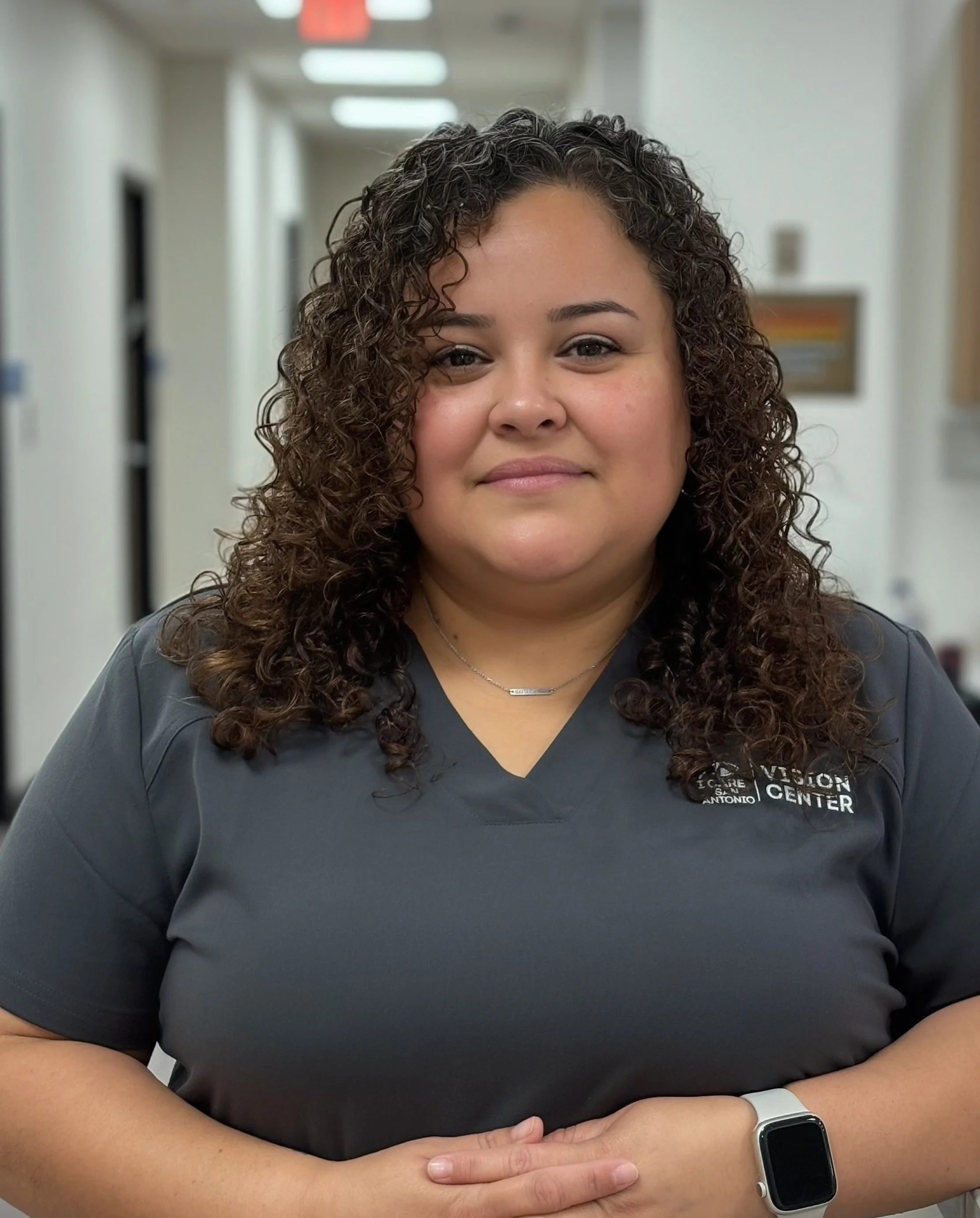 A woman with curly hair wearing a gray scrub top and a smartwatch, standing in a hallway of a medical or care center.
