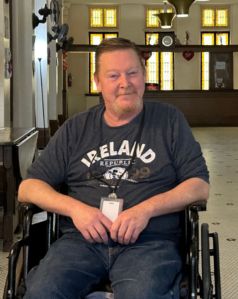 A man sitting in a wheelchair inside a church with stained glass windows, wearing a dark T-shirt with the word 'Ireland' on it and smiling.
