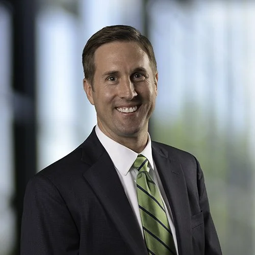 Professional headshot of a man in a suit with a smile, standing in front of a blurred office background.