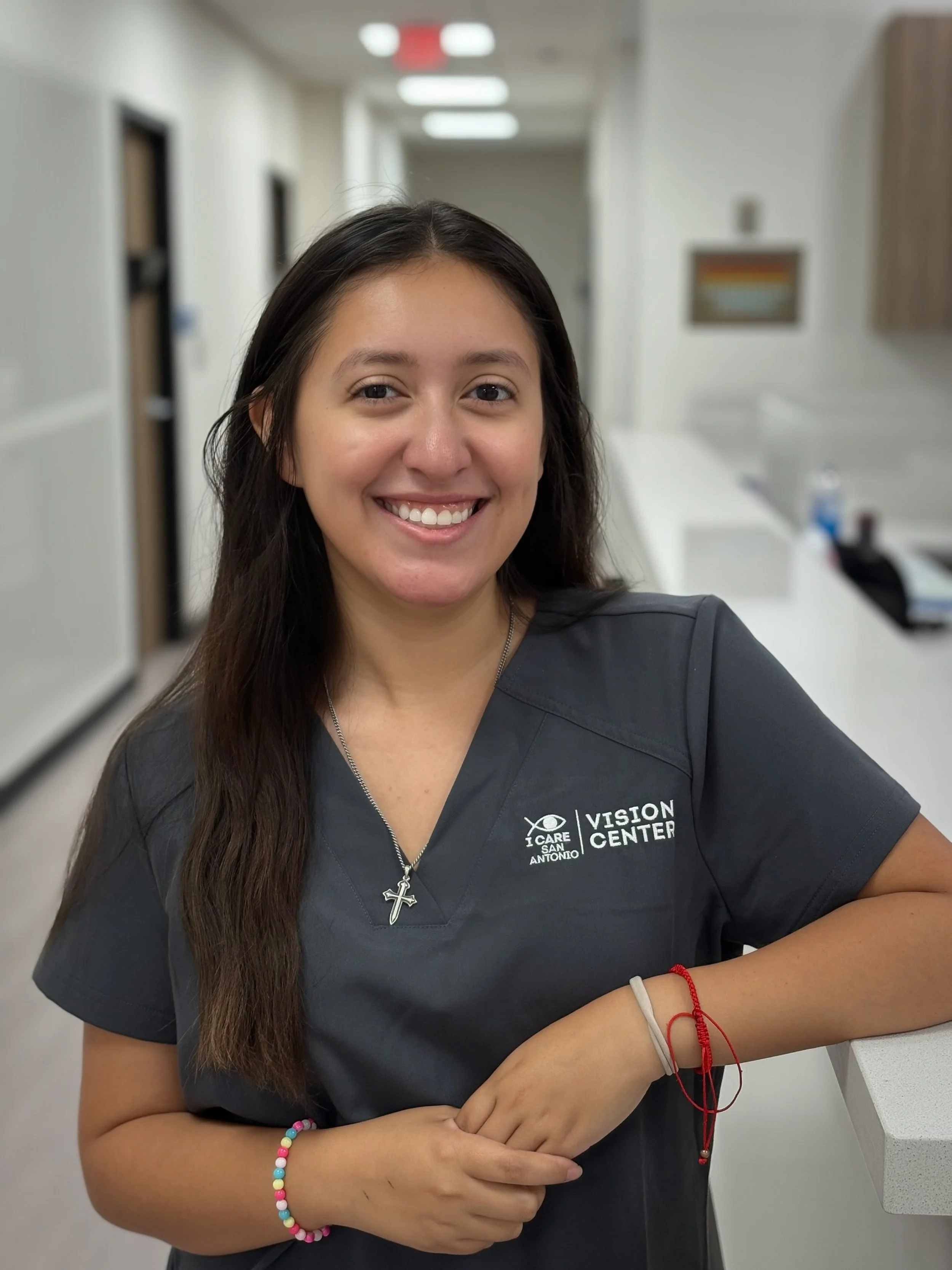 Smiling woman in grey scrubs with 'Vision Center' and 'I Care San Antonio' logos, standing in a clinical setting, wearing a cross necklace and colorful bracelets.