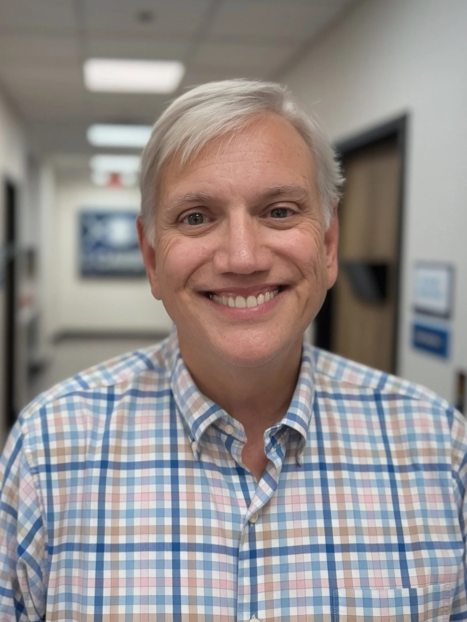 A smiling middle-aged man with gray hair in a checkered shirt standing in an indoor corridor.